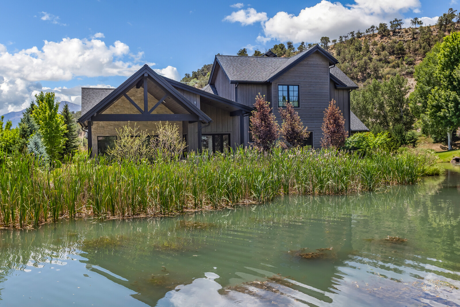 This stunning custom home features a modern farmhouse aesthetic with dark wood siding, stone accents, and a prominent timber-framed covered patio. The residence is beautifully situated behind a tranquil pond filled with tall reeds, creating a serene and picturesque setting. The composition captures the home from a low-angle perspective across the water, emphasizing its integration with the surrounding natural landscape and mountain backdrop.