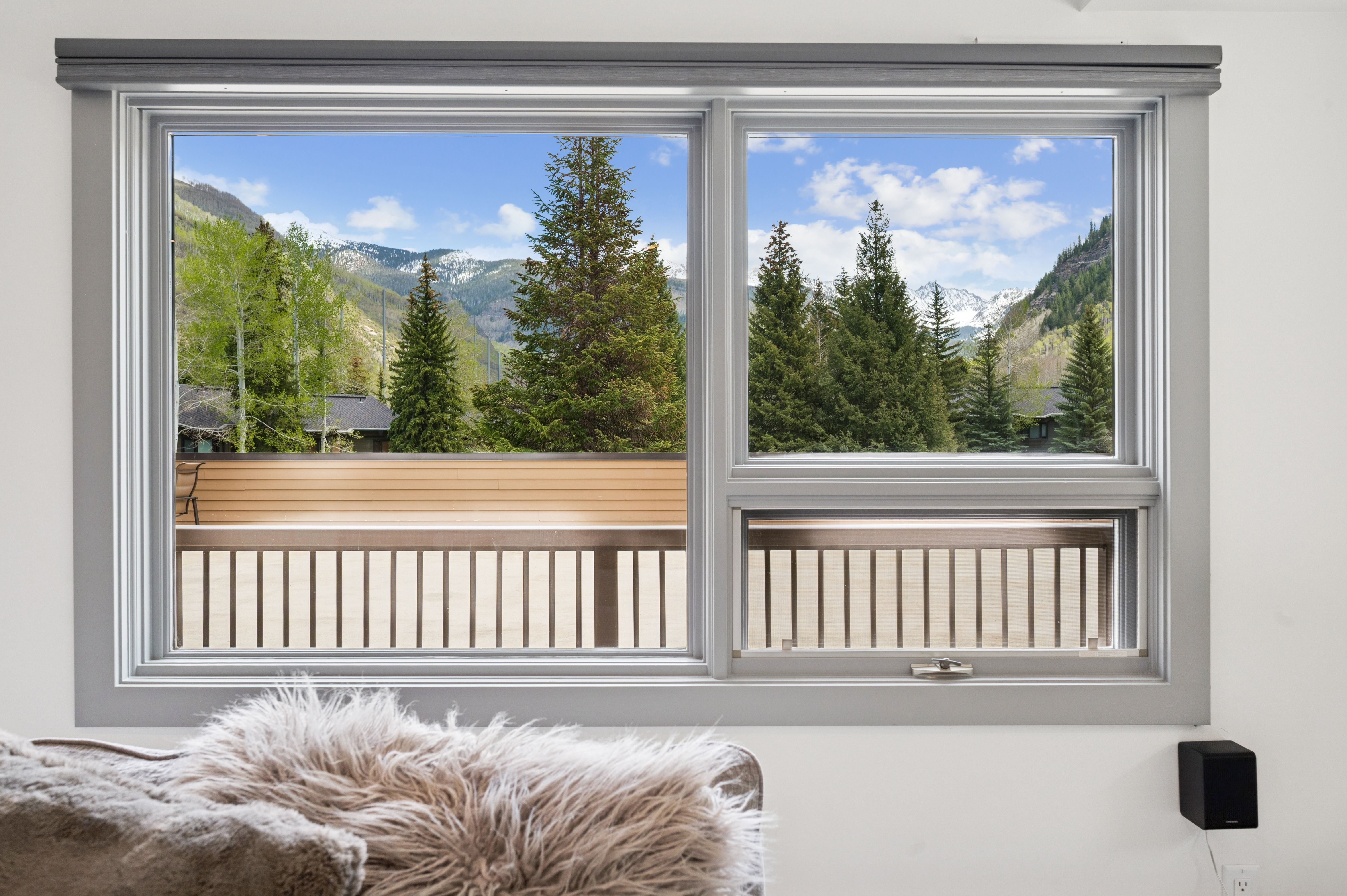This interior shot captures a cozy living space focused on a large, multi-pane window that frames a breathtaking mountain and forest view. In the foreground, a plush, textured throw pillow adds a sense of comfort and warmth to the room. The clean, neutral wall and modern window frame highlight the scenic outdoor landscape, creating a serene and inviting atmosphere.