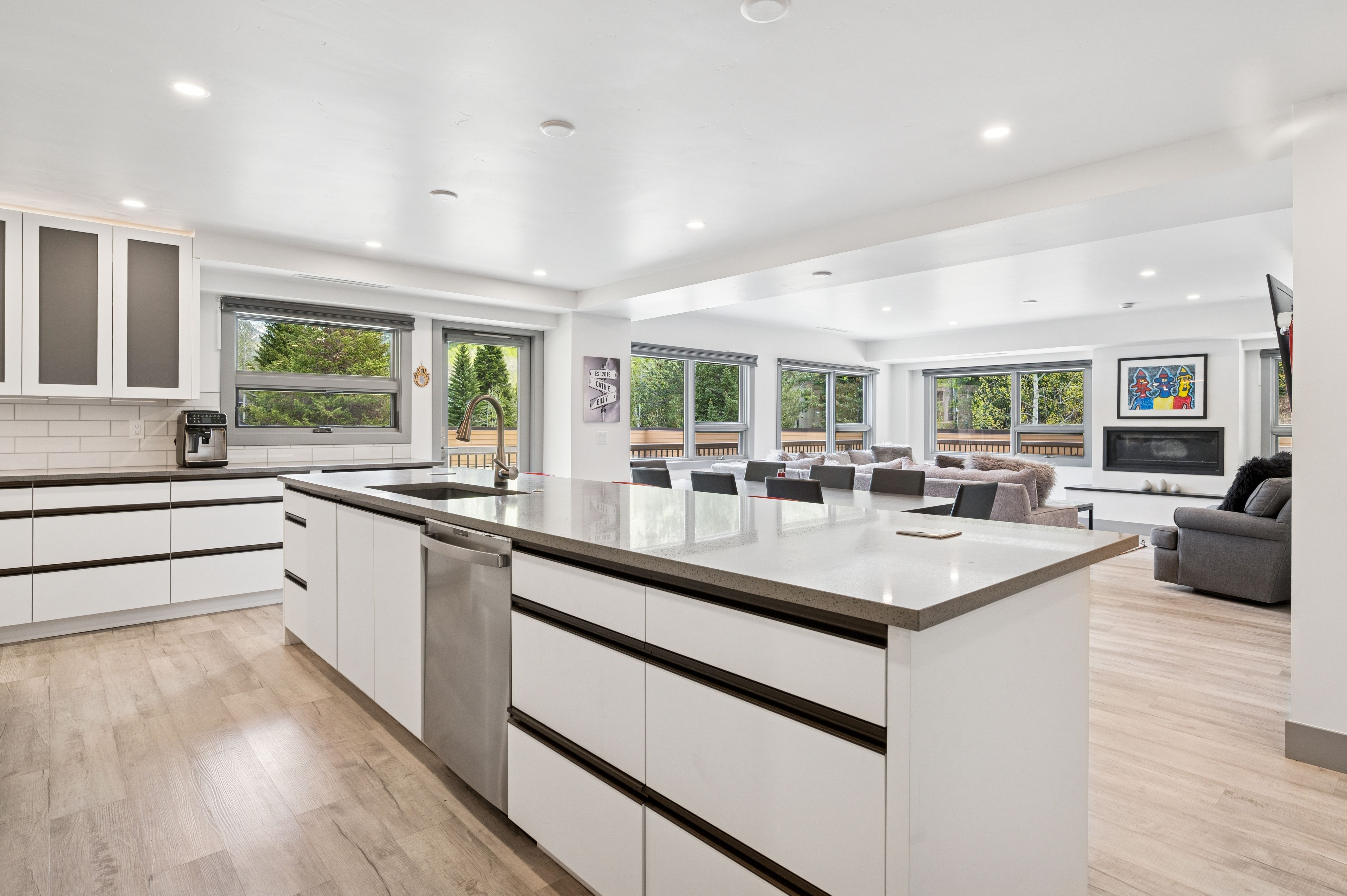 This bright and modern open-concept kitchen features sleek white cabinetry, a large central island with a stainless steel dishwasher, and light wood-look flooring. The space flows seamlessly into a spacious living area with a fireplace, creating an airy and inviting atmosphere perfect for entertaining. Large windows throughout the room provide ample natural light and views of the surrounding greenery.