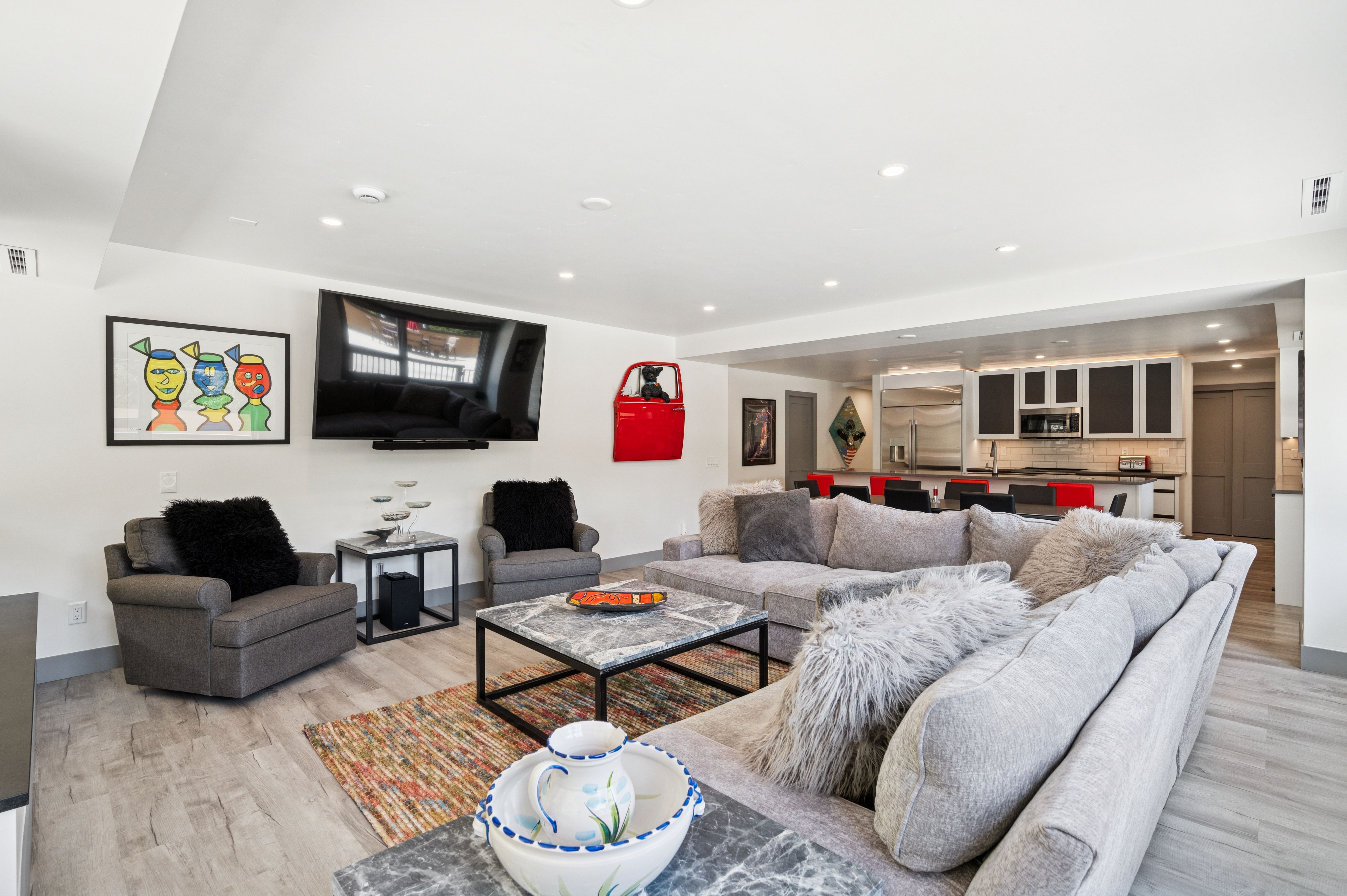 This spacious, open-concept living room features a large grey sectional sofa, two matching armchairs, and a marble-topped coffee table set atop a colorful area rug. The room is anchored by a wall-mounted television and accented with unique decor, including a red car door piece and vibrant abstract art. The perspective captures the flow from the cozy seating area into the adjacent kitchen, highlighting a bright, modern, and inviting living space.