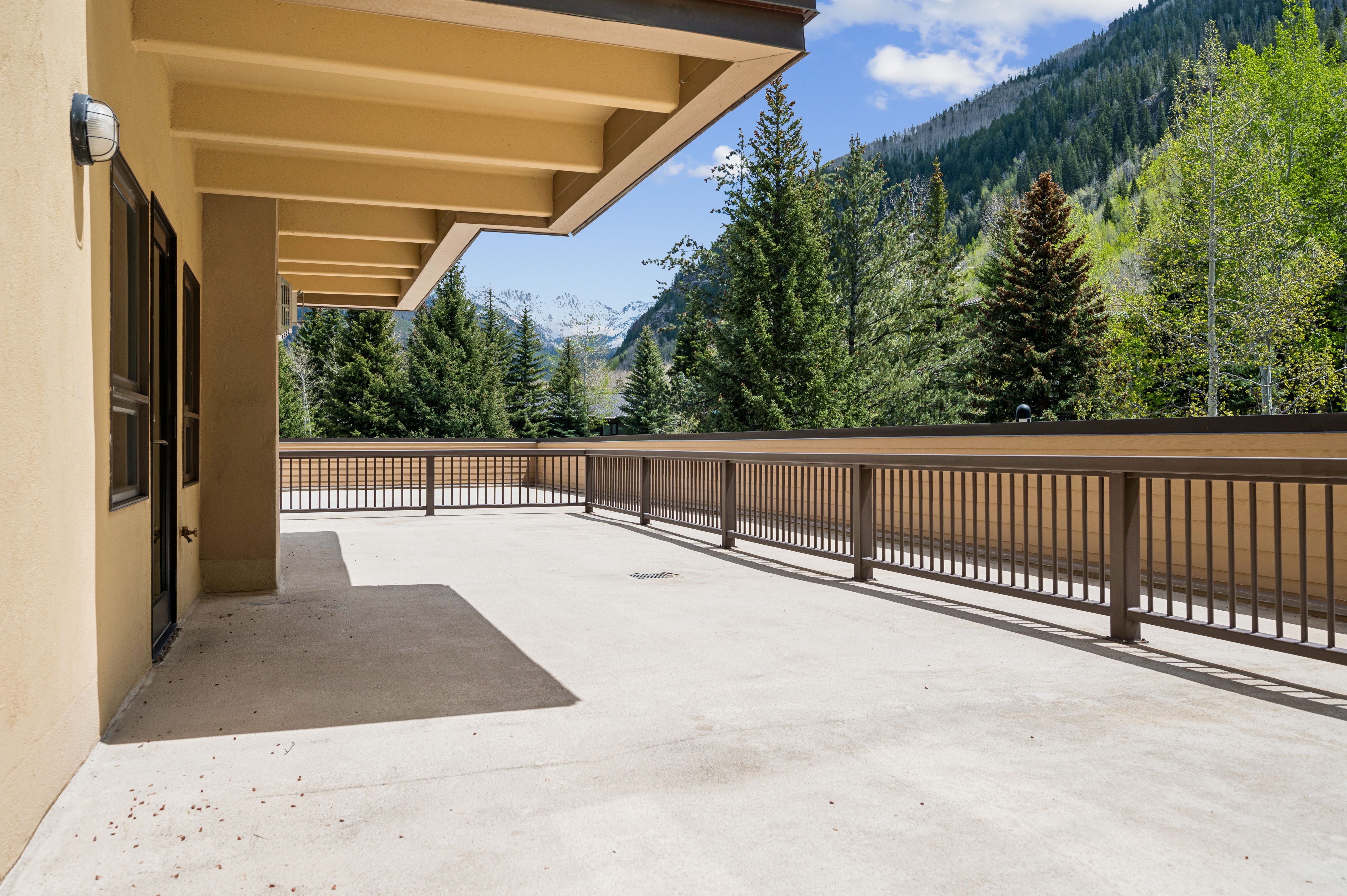 This spacious, covered balcony offers a stunning panoramic view of snow-capped mountains and a lush evergreen forest. The area features a clean concrete floor, a sturdy metal railing, and a neutral-toned exterior wall, creating a serene and inviting outdoor living space. The perspective captures the expansive nature of the deck, emphasizing its potential for relaxation and scenic enjoyment.