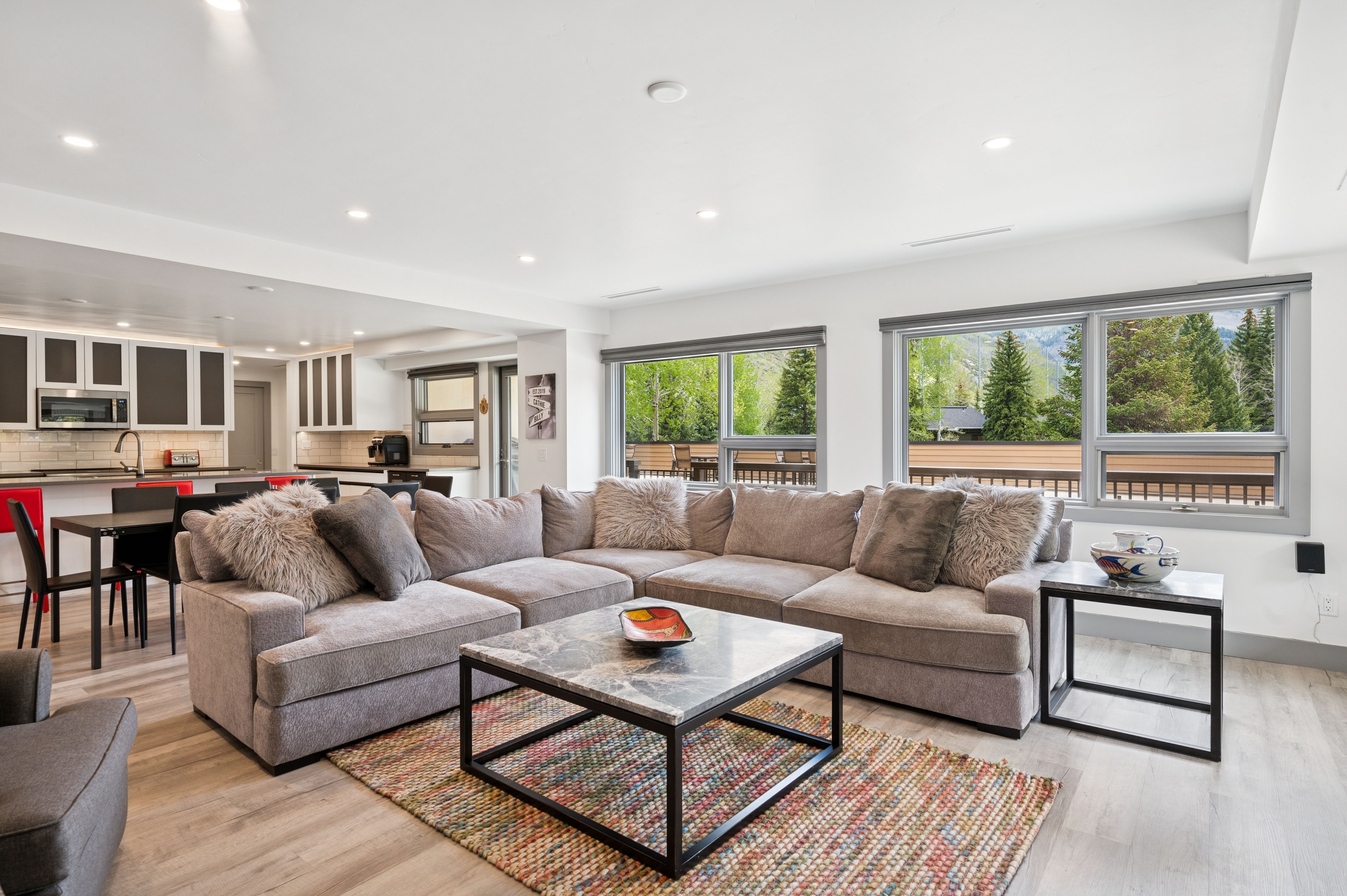 This spacious, open-concept living room features a large, plush grey sectional sofa centered around a modern marble-topped coffee table with a black metal frame. The room is bathed in natural light from large windows that offer a scenic view of the surrounding trees and mountains, while the adjacent kitchen area maintains a cohesive, contemporary aesthetic. The light wood-look flooring and neutral color palette create an inviting and airy atmosphere perfect for relaxation or entertaining.