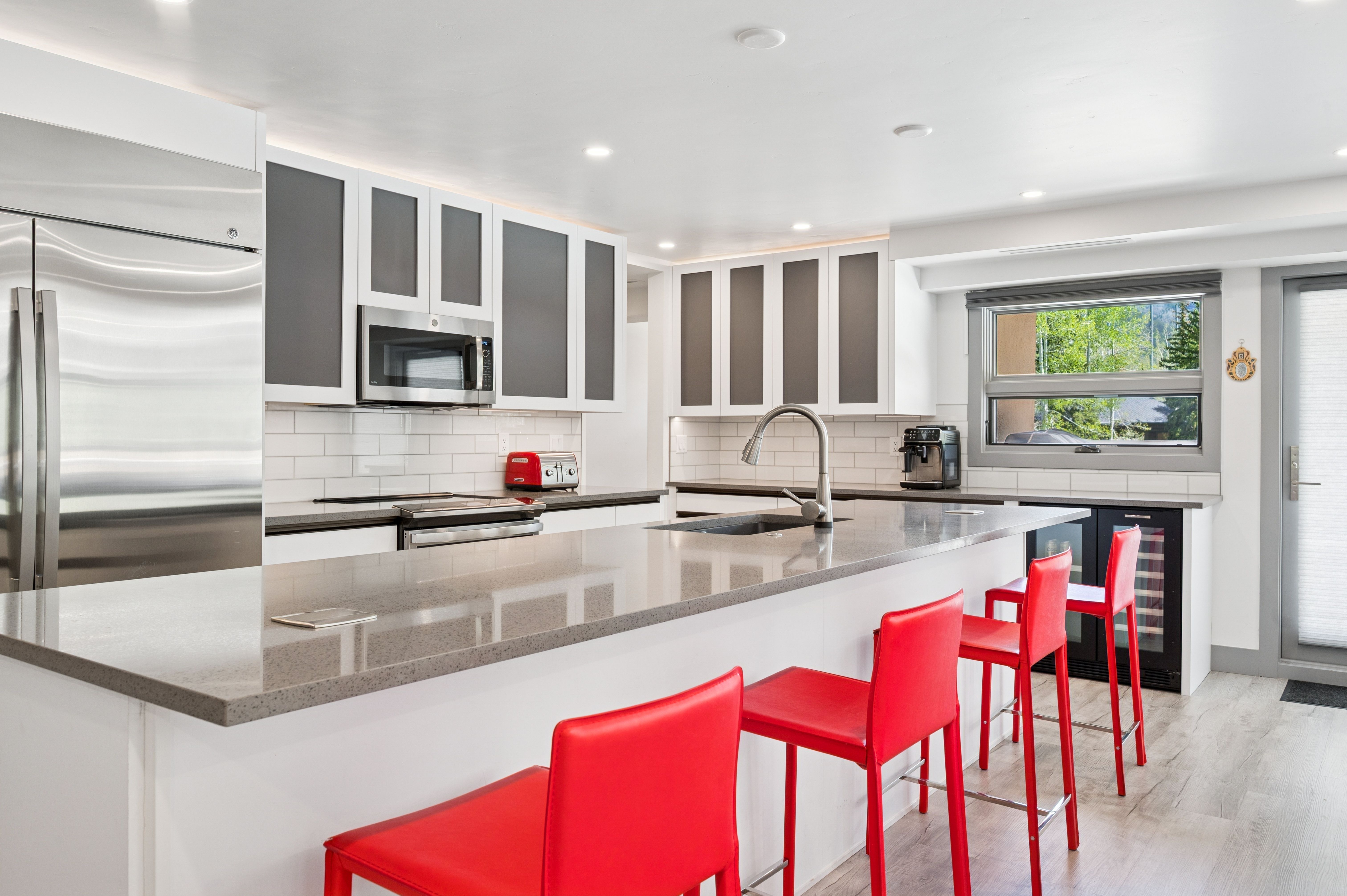This modern, high-contrast kitchen features sleek white cabinetry with dark grey accents, stainless steel appliances, and a spacious quartz-topped island. Three vibrant red bar stools provide a bold pop of color against the neutral palette, while recessed lighting and a subway tile backsplash complete the contemporary aesthetic. The perspective is a wide-angle shot from the kitchen island, showcasing the open-concept layout and the seamless transition to the outdoor view through the window.