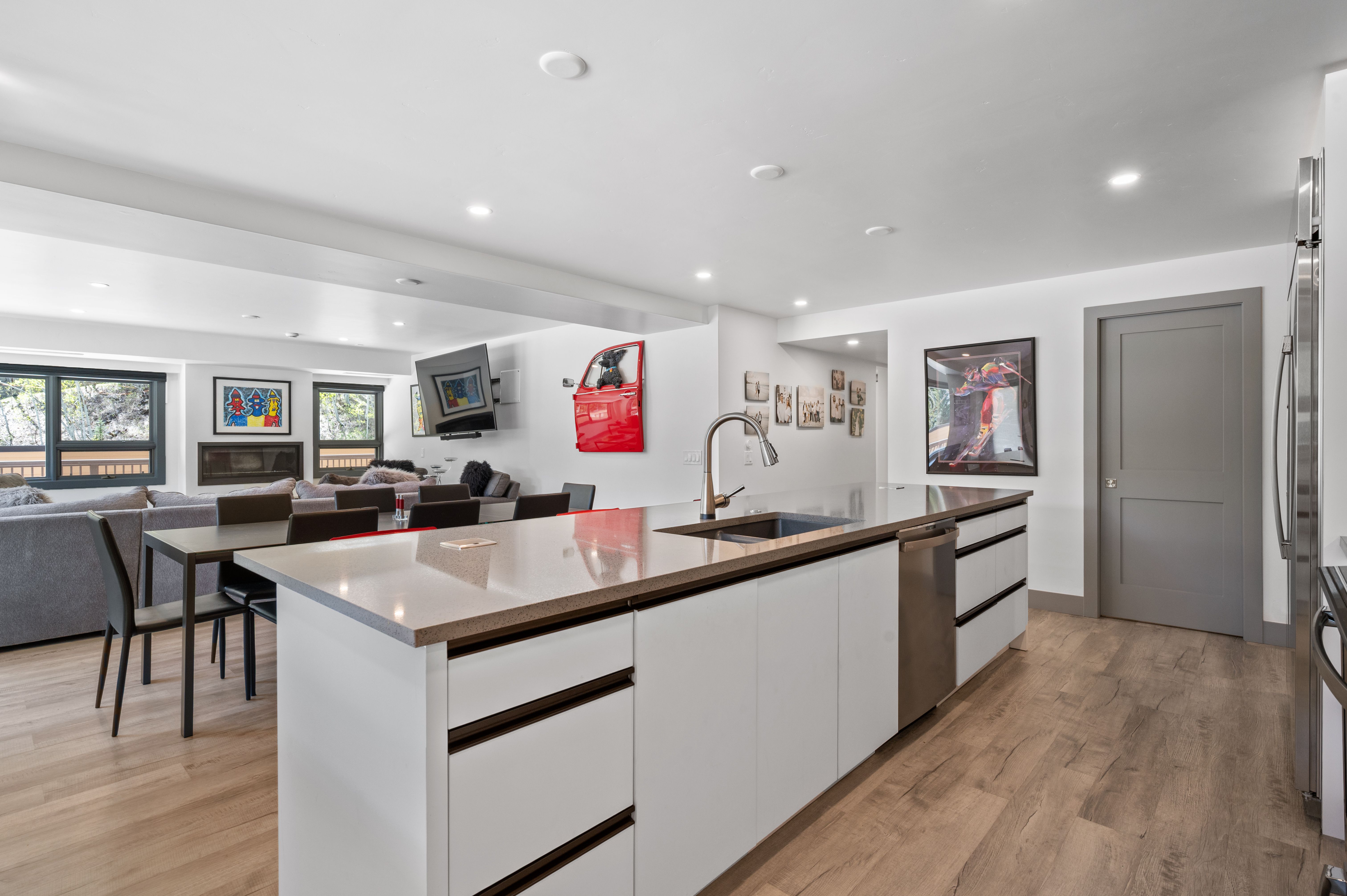 This modern, open-concept kitchen features a large central island with a sleek quartz countertop, stainless steel sink, and integrated dishwasher. The space is bright and airy, flowing seamlessly into a dining and living area decorated with contemporary art and a unique red car door wall accent. The light wood flooring and minimalist white cabinetry create a clean, sophisticated aesthetic perfect for modern living.