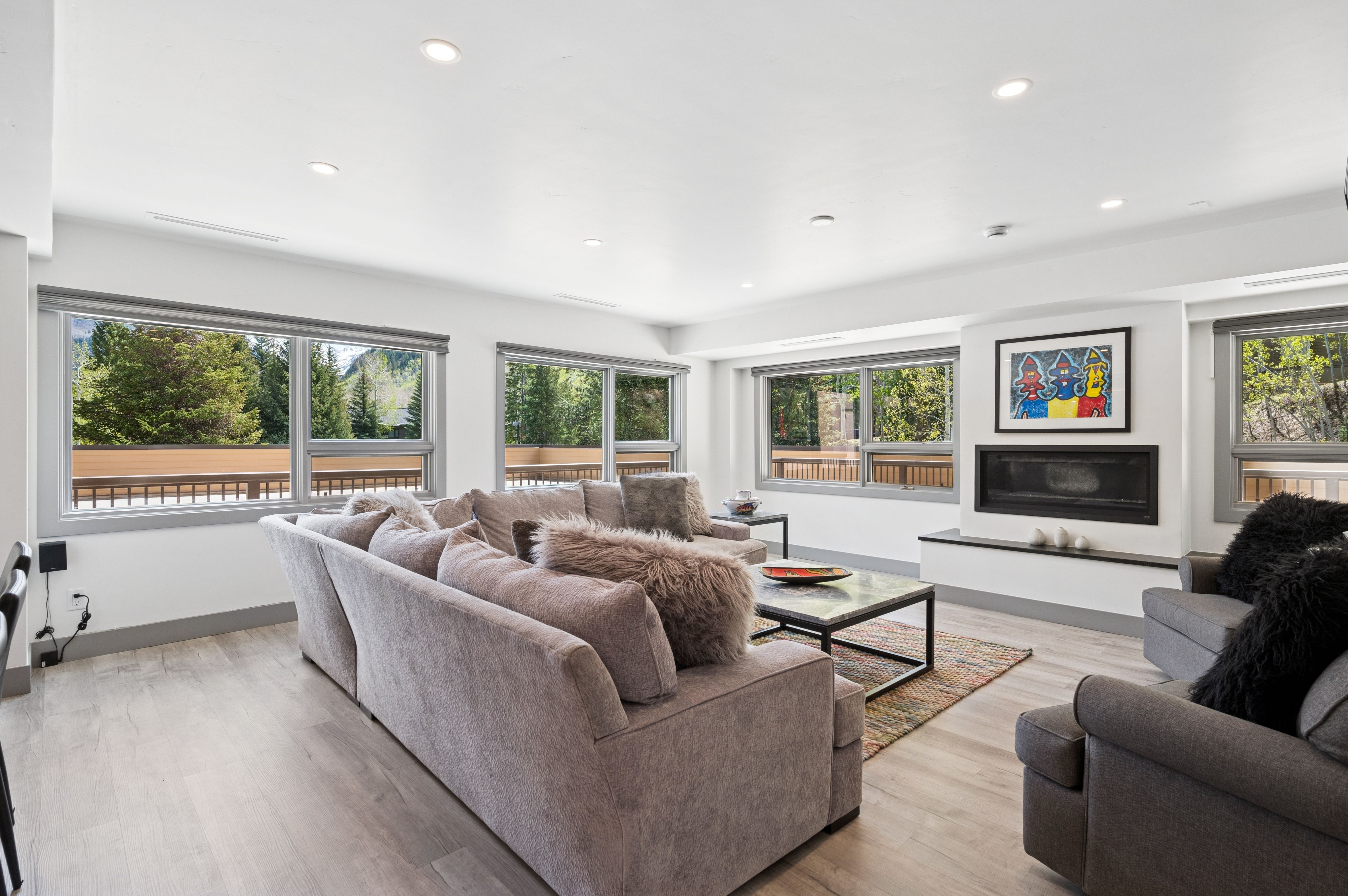 This bright and spacious living room features a large, comfortable sectional sofa with plush, textured pillows and a modern coffee table set on a patterned area rug. A sleek, built-in fireplace serves as a focal point, adorned with colorful abstract artwork above it, while expansive windows offer scenic views of the surrounding trees and mountains. The room is finished with light wood-look flooring and recessed lighting, creating a warm and inviting atmosphere.