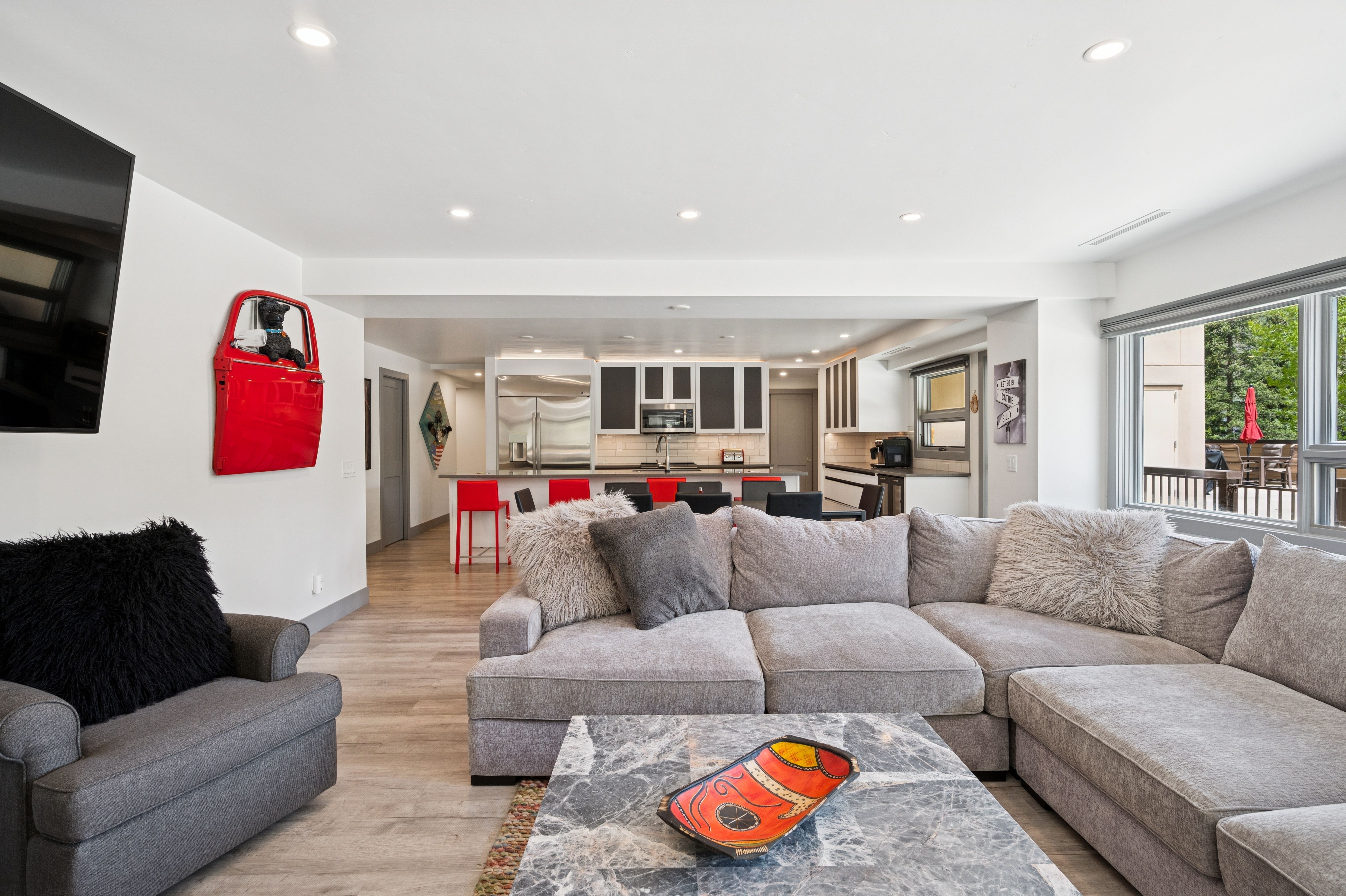This open-concept living space features a large, plush grey sectional sofa and a matching armchair, centered around a sophisticated marble-topped coffee table. The room flows seamlessly into a modern kitchen with a breakfast bar, creating an inviting and airy atmosphere perfect for entertaining. Notable decor includes a vibrant red car door wall art piece and a large television, blending contemporary comfort with unique, eclectic style.