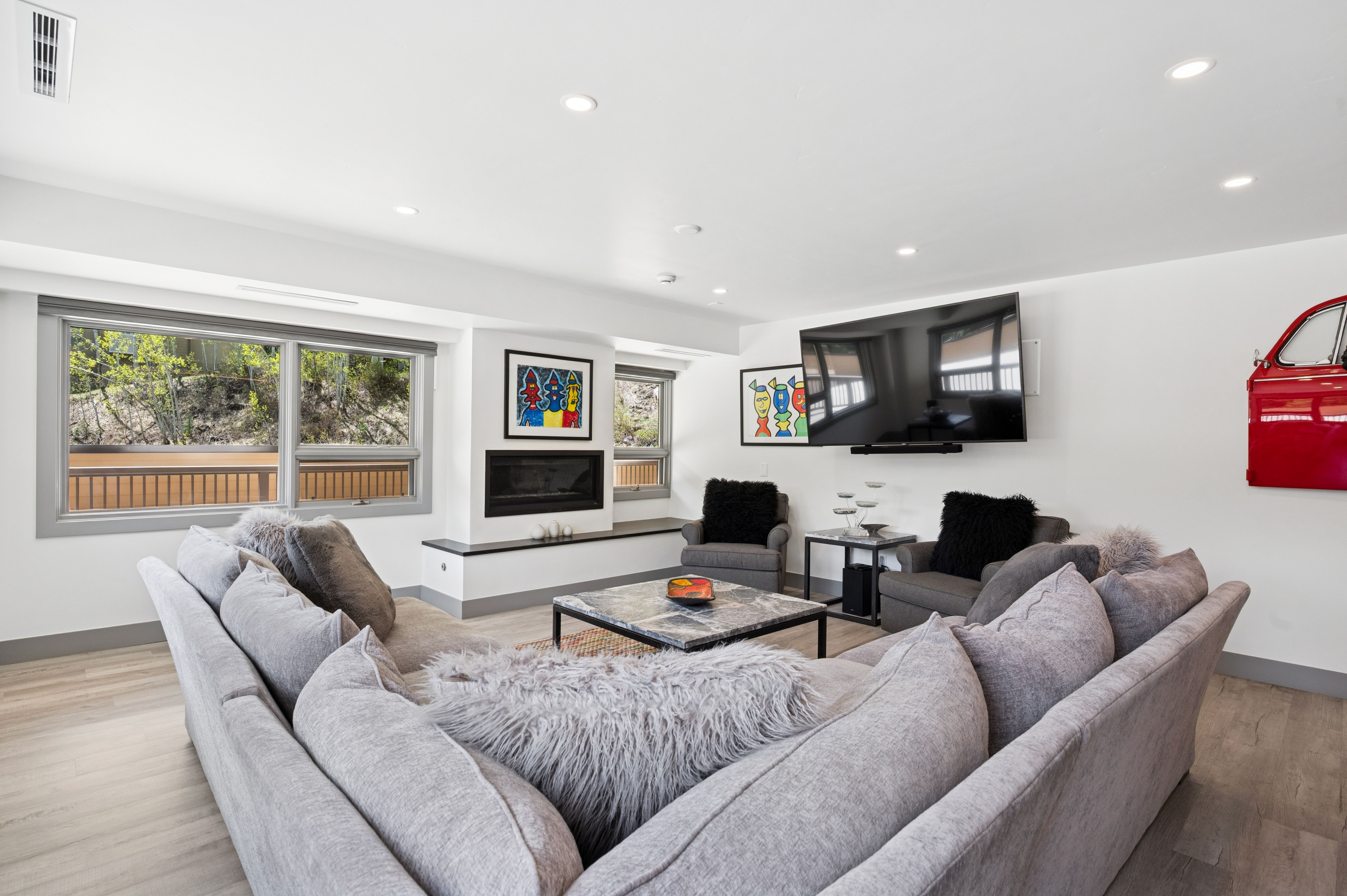 This contemporary living room features a large, plush grey sectional sofa arranged around a marble-topped coffee table, creating a cozy and inviting atmosphere. The space is accented by vibrant, abstract artwork, a sleek fireplace, and a unique red automotive-themed wall decor piece. Large windows provide natural light and a view of the outdoor surroundings, while the neutral color palette and soft textures offer a modern, comfortable aesthetic.