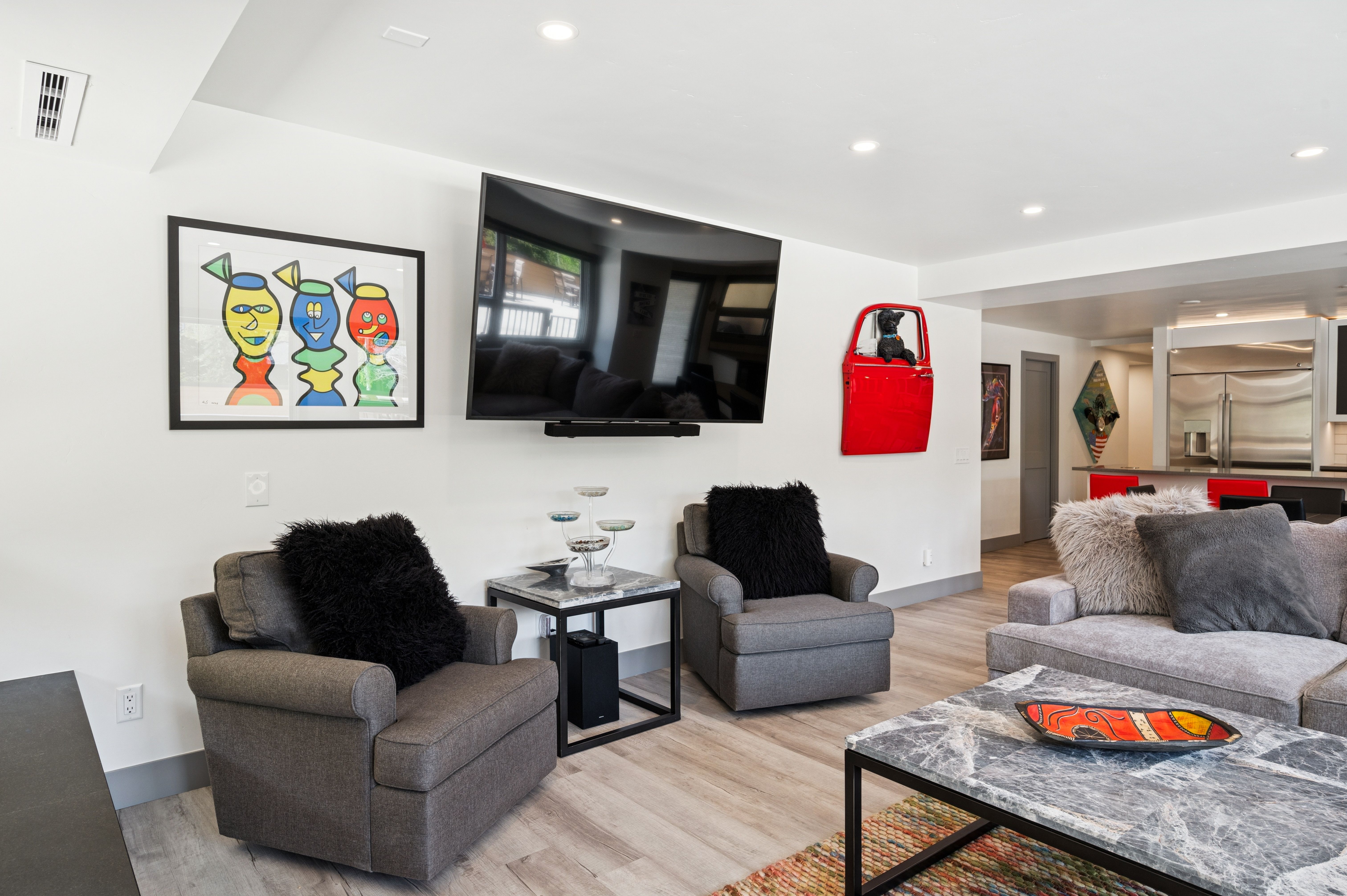 This contemporary living room features a comfortable seating area with two grey armchairs and a plush sofa, centered around a marble-topped coffee table. The space is accented by vibrant, eclectic wall art and a unique red car door wall hanging, creating a playful and modern aesthetic. The open-concept layout flows seamlessly into a bright kitchen area in the background, highlighting the home's spacious and inviting atmosphere.