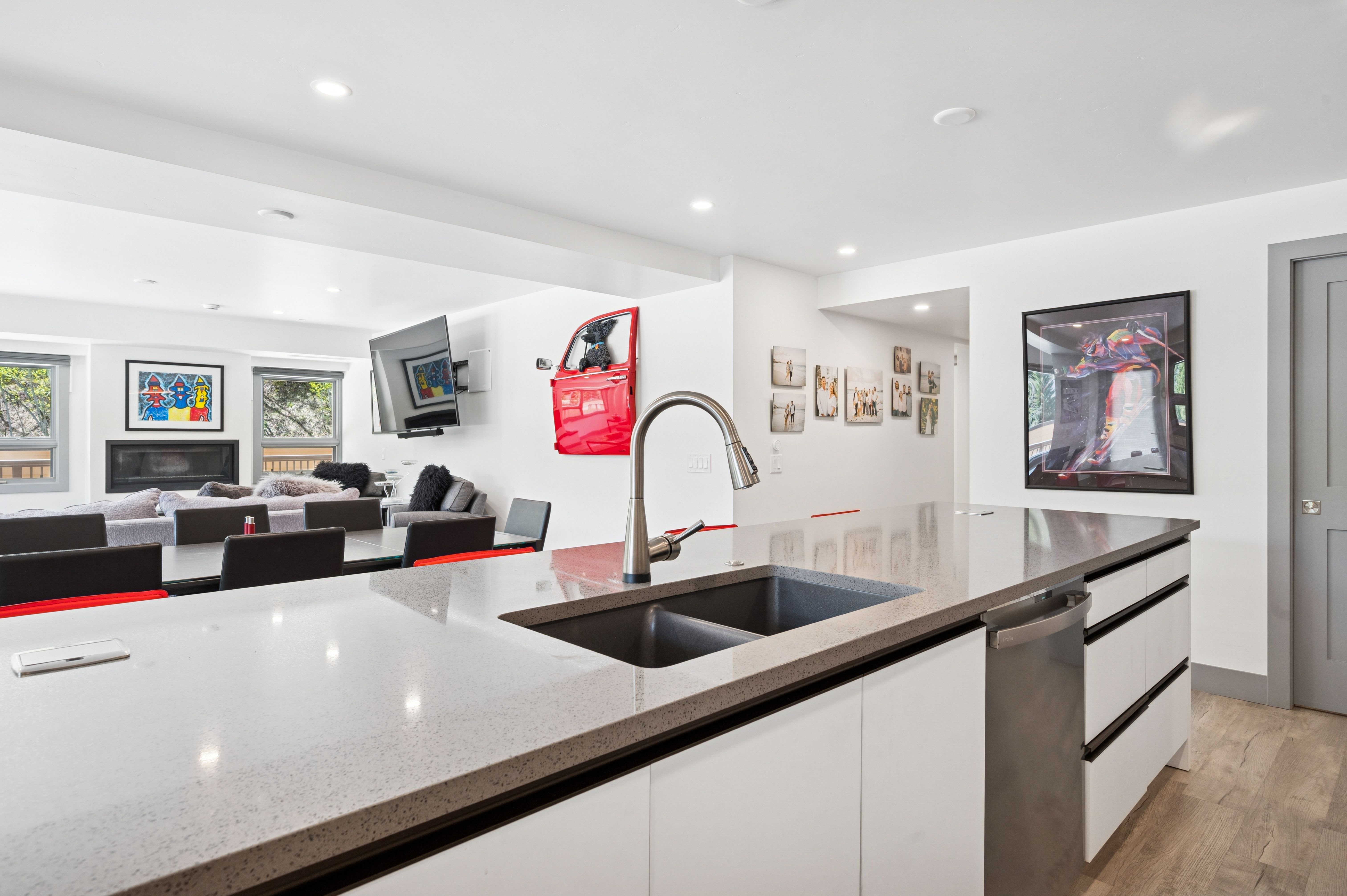 This modern kitchen features a sleek, minimalist design with white cabinetry, a dark grey quartz countertop, and a double-basin undermount sink. The open-concept layout seamlessly connects the kitchen to the dining and living areas, which are visible in the background, creating a spacious and airy atmosphere. The space is accented by contemporary artwork and a unique red car door wall decoration, adding a vibrant, personalized touch to the clean, neutral aesthetic.