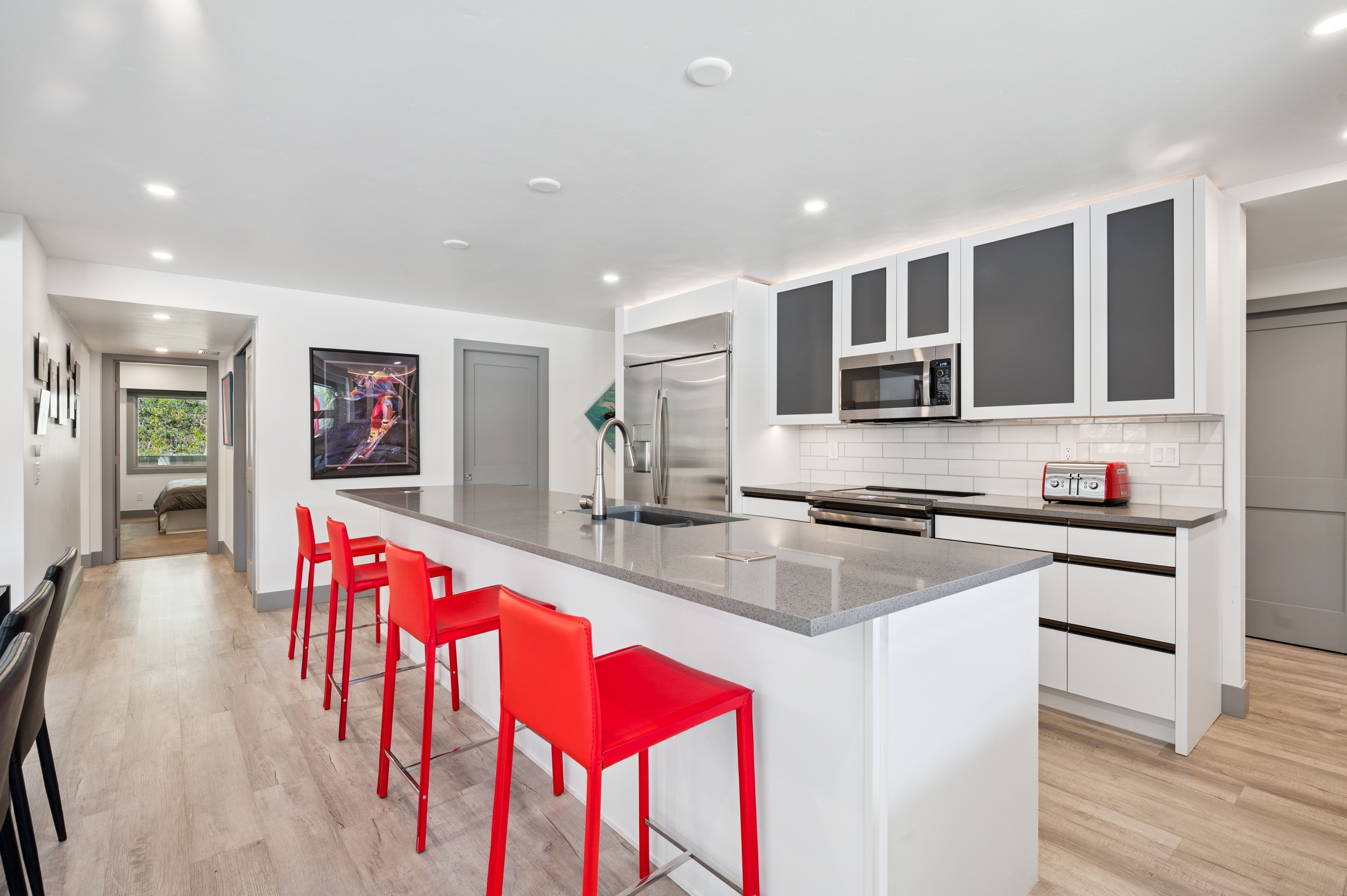 This modern, open-concept kitchen features a large central island with a gray quartz countertop and four vibrant red bar stools, creating a striking focal point. The space is equipped with sleek white cabinetry, stainless steel appliances, and a subway tile backsplash, all illuminated by recessed lighting. The light wood-look flooring and neutral color palette provide a clean, contemporary aesthetic that flows seamlessly into the adjacent hallway.