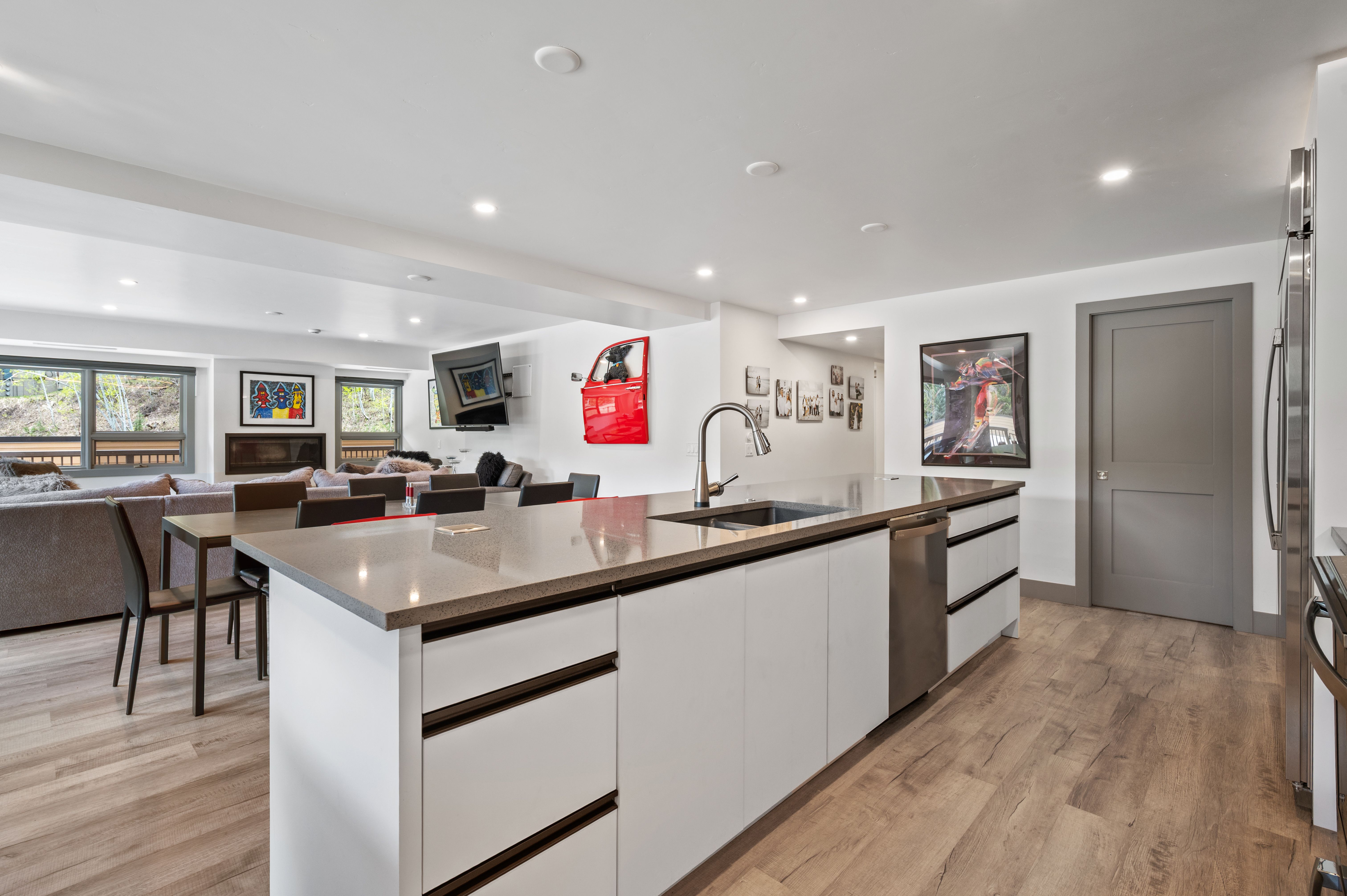 This modern, open-concept kitchen features a large central island with a sleek gray countertop and white cabinetry, seamlessly flowing into a dining and living area. The space is brightened by recessed lighting and light wood-tone flooring, creating a clean and contemporary aesthetic. A unique red car door wall art piece adds a vibrant pop of color, while the perspective highlights the functional layout and connection between the kitchen and the rest of the home.