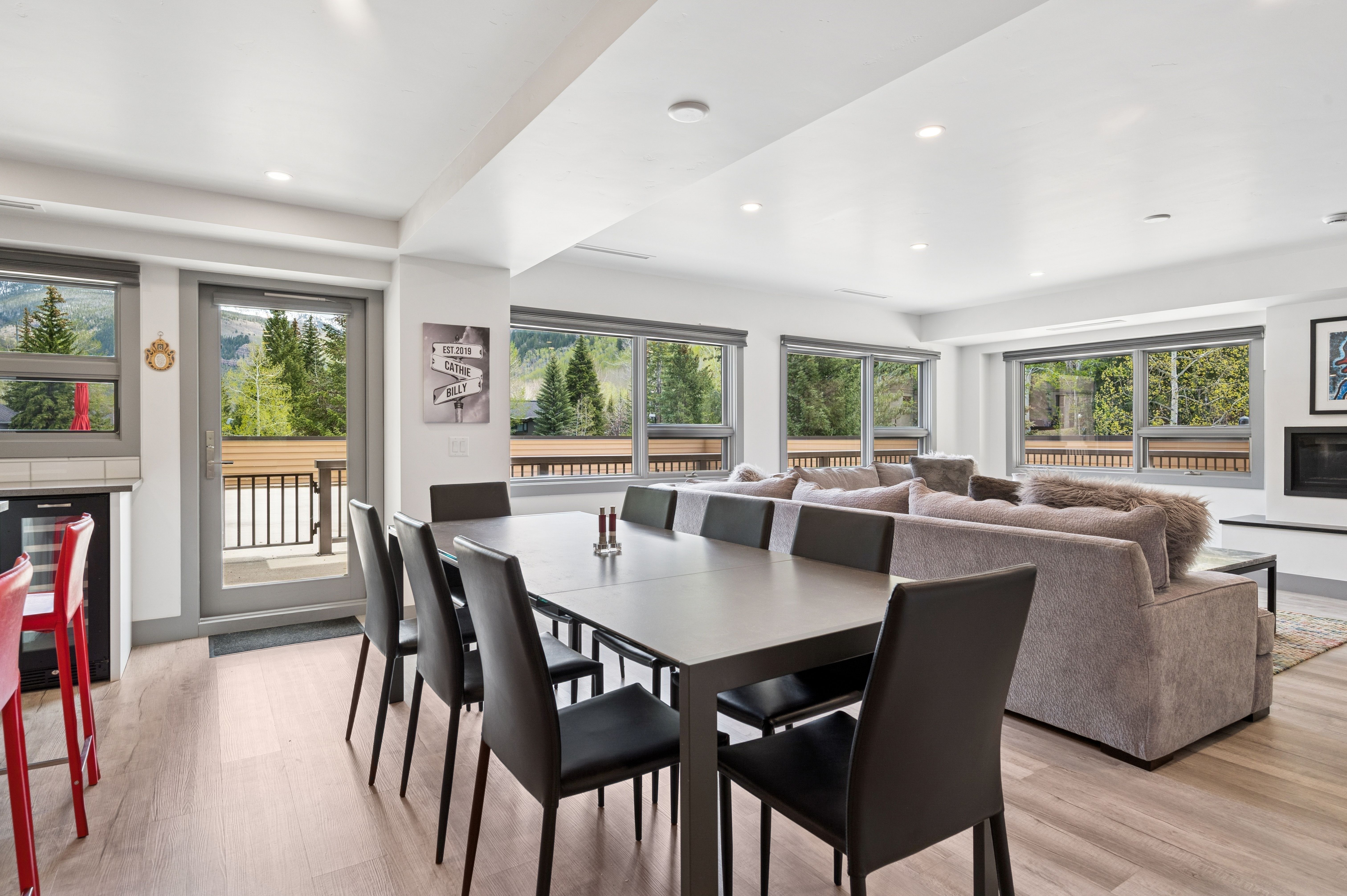 This open-concept living and dining area features a sleek, dark dining table surrounded by modern black chairs, seamlessly transitioning into a cozy living space with a large, plush sectional sofa. The room is bathed in natural light from expansive windows that offer scenic views of the surrounding forest and mountains. The neutral color palette, light wood flooring, and clean lines create a contemporary and inviting atmosphere perfect for entertaining.