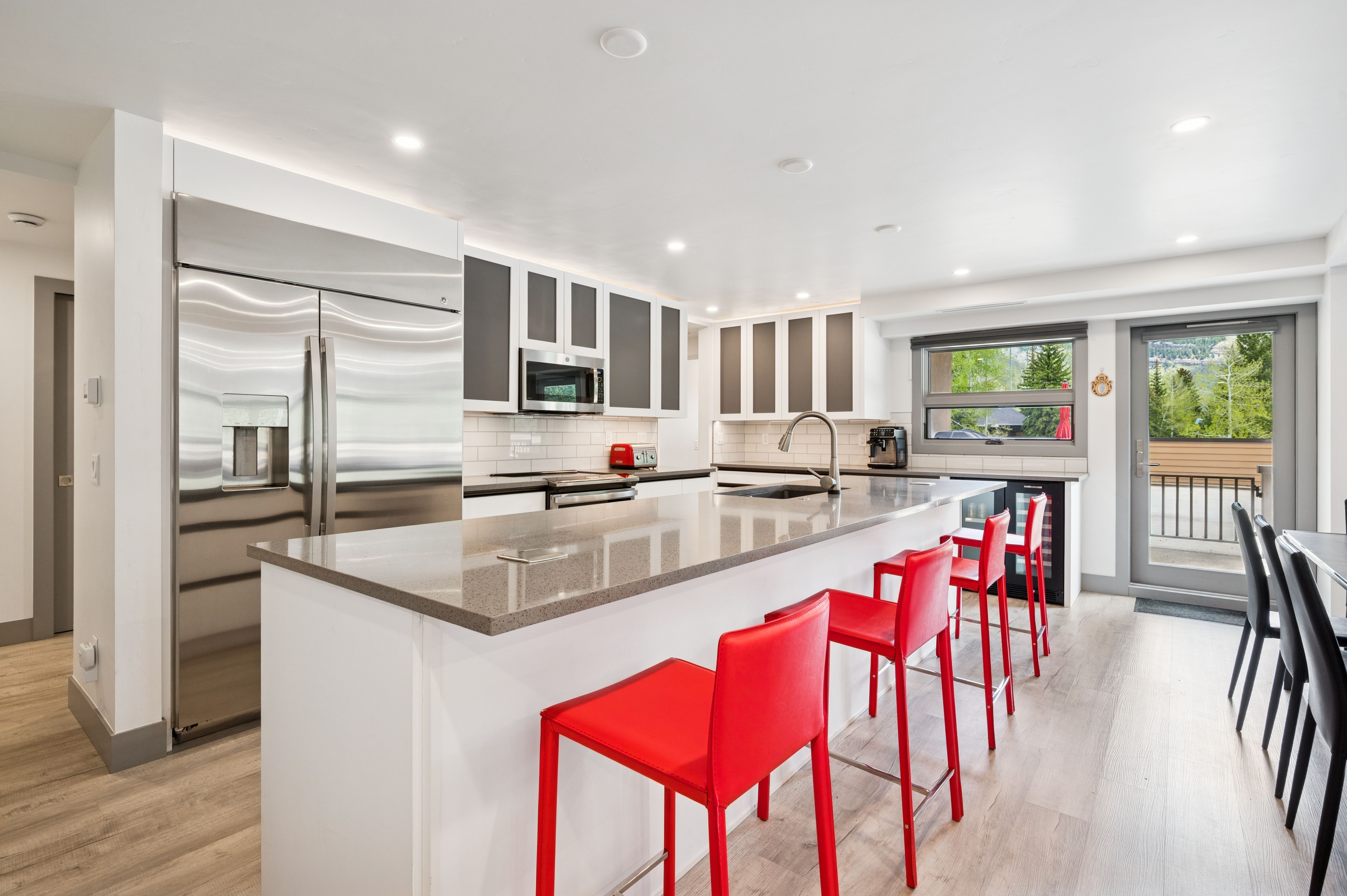 This modern, open-concept kitchen features sleek white cabinetry, stainless steel appliances, and a spacious island topped with a grey stone countertop. Vibrant red bar stools provide a bold pop of color against the neutral palette, while a large window and glass door invite natural light and offer a view of the outdoors. The space is clean, contemporary, and well-lit with recessed lighting, creating an inviting atmosphere for cooking and entertaining.