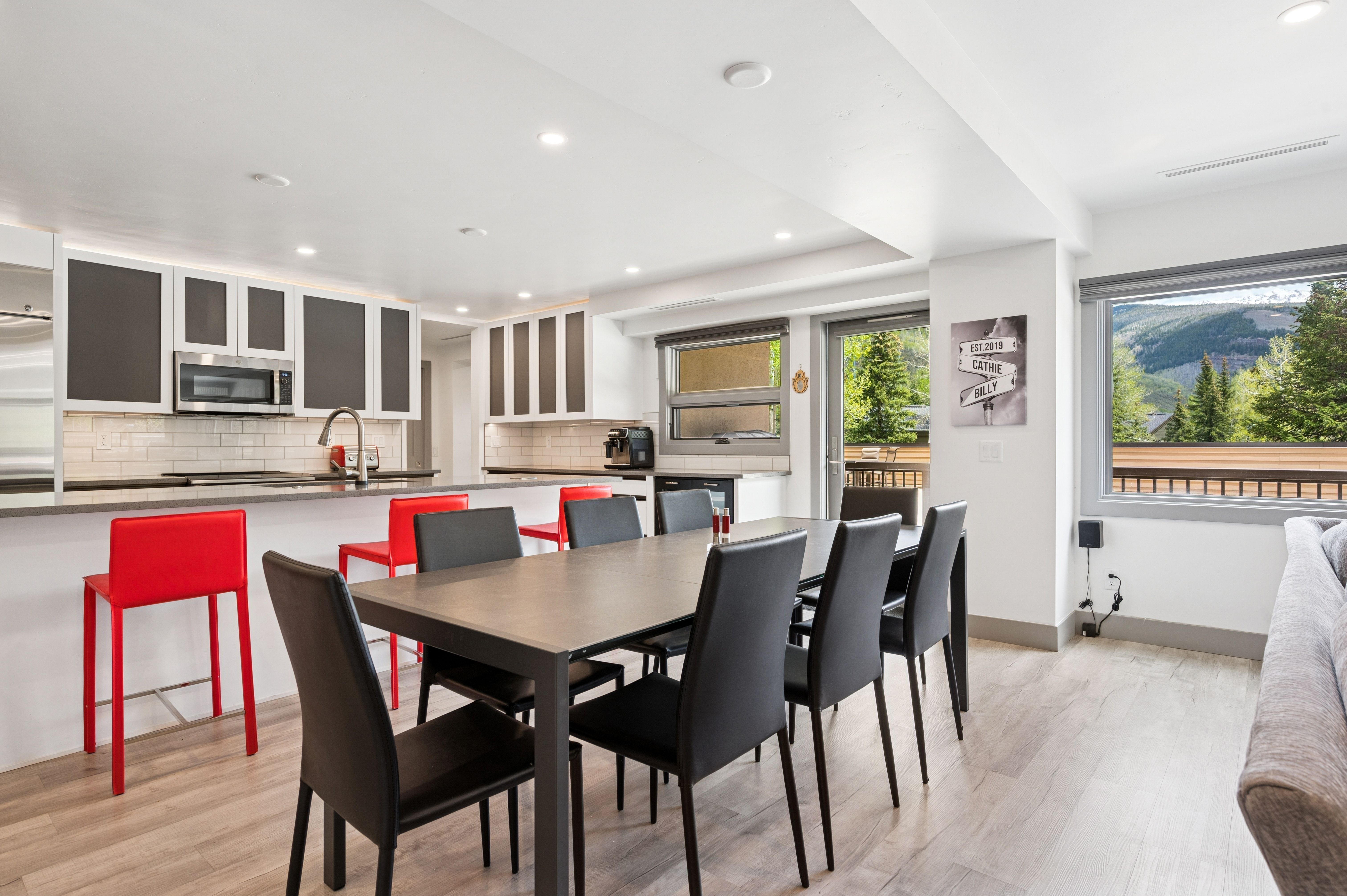 This open-concept space seamlessly blends a modern kitchen with a dining area, featuring sleek white cabinetry, stainless steel appliances, and a breakfast bar with vibrant red stools. A large dark wood dining table with matching chairs sits in the foreground, while a sliding glass door provides natural light and a view of the surrounding mountain landscape. The room is characterized by its clean, contemporary aesthetic, light wood-look flooring, and a bright, airy atmosphere.