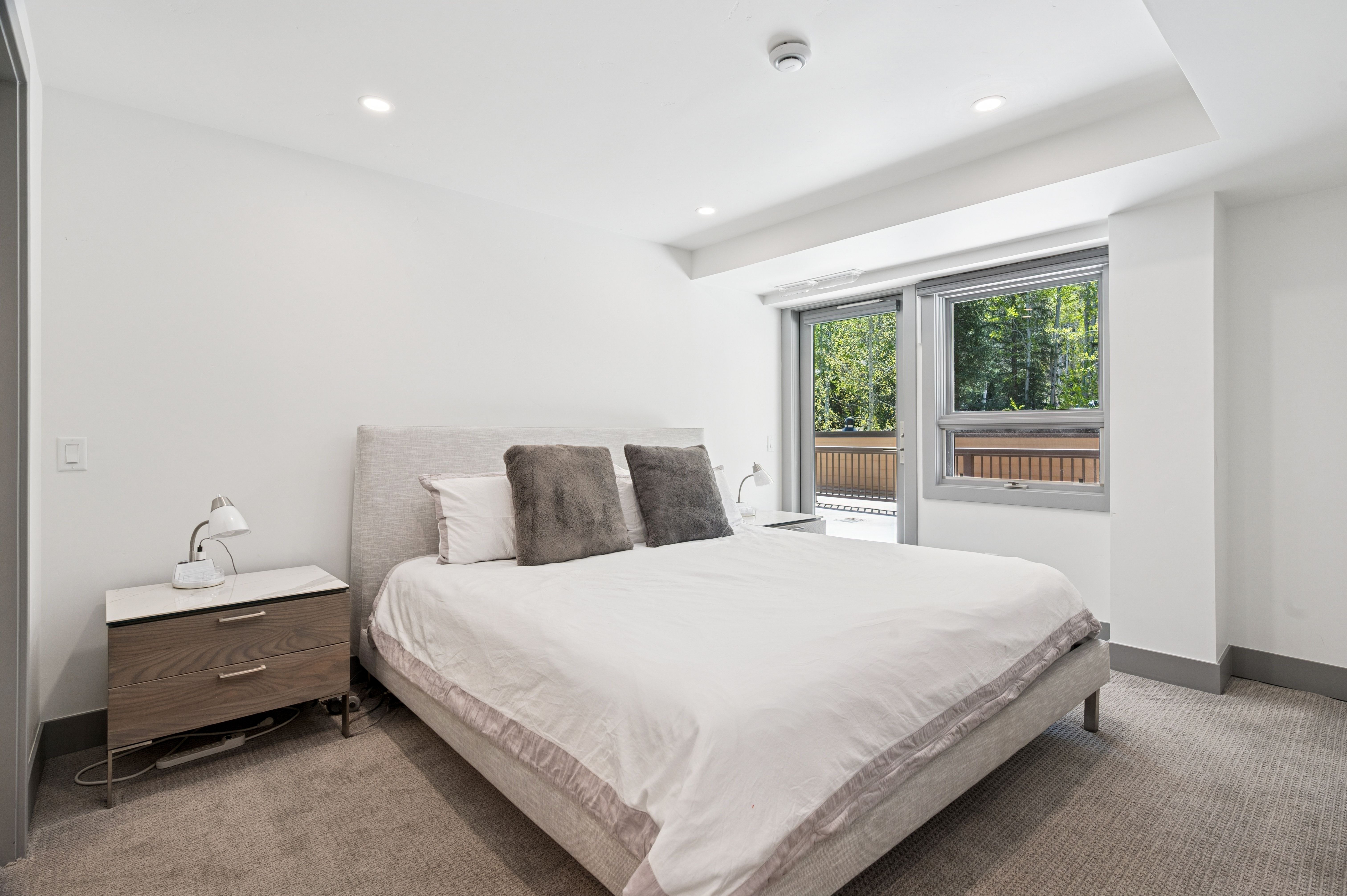 This bright and airy primary bedroom features a neutral color palette, a plush upholstered bed frame, and a modern wood-toned nightstand with a marble top. Large windows and a glass door provide ample natural light and a view of an outdoor balcony, creating a serene and inviting atmosphere. The room is finished with soft carpeting and minimalist decor, emphasizing a clean, contemporary aesthetic.