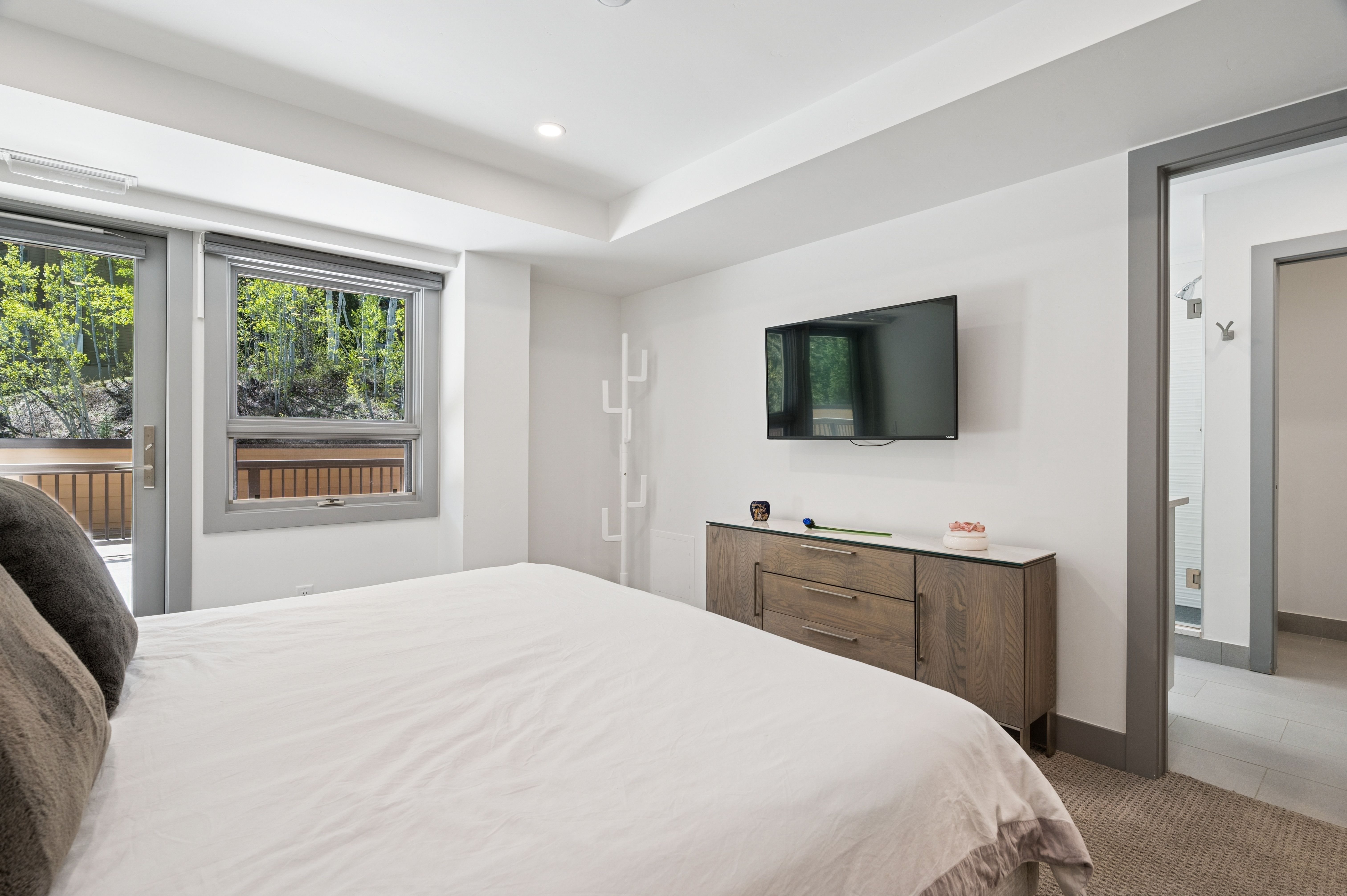 This modern primary bedroom features a clean, minimalist aesthetic with a neutral color palette and plush carpeting. A large window and glass door provide ample natural light and a view of the surrounding trees, while a wall-mounted television sits above a sleek wooden dresser. The room flows seamlessly into an adjacent bathroom, creating a bright and inviting atmosphere.