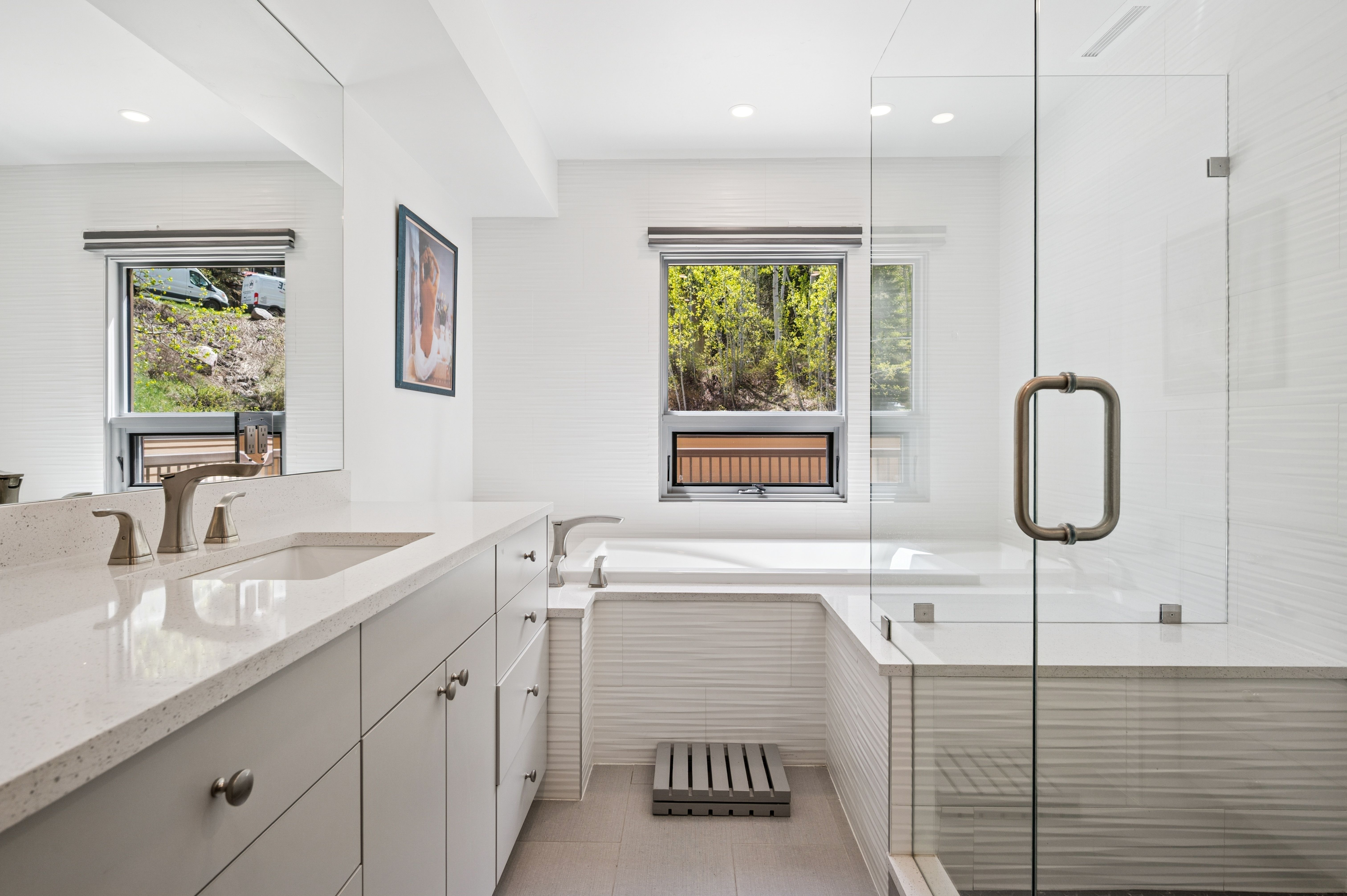 This modern primary bathroom features a clean, minimalist aesthetic with white textured wall tiles, a sleek vanity with quartz countertops, and a spacious glass-enclosed shower. A deep soaking tub is positioned beneath a window that provides natural light and a view of the surrounding greenery, creating a serene and spa-like atmosphere. The neutral color palette and high-end finishes contribute to a bright, airy, and luxurious feel.