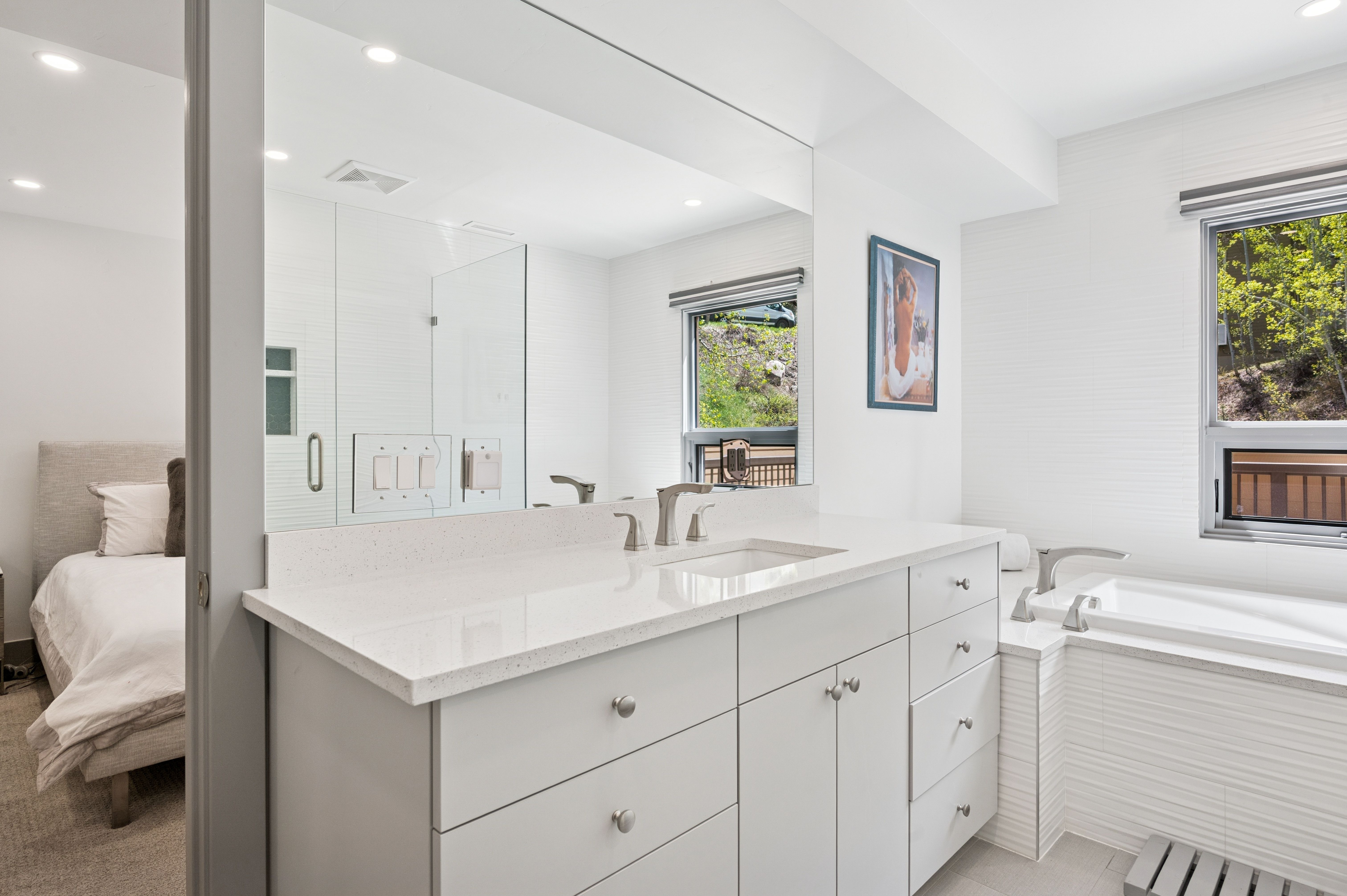 This bright and modern primary bathroom features a sleek white vanity with a quartz countertop, a large wall-to-wall mirror, and a deep soaking tub positioned under a window. The space is finished with clean, textured white wall tiles and offers a seamless transition into the adjacent bedroom. The overall aesthetic is minimalist and serene, emphasizing a clean, spa-like atmosphere.