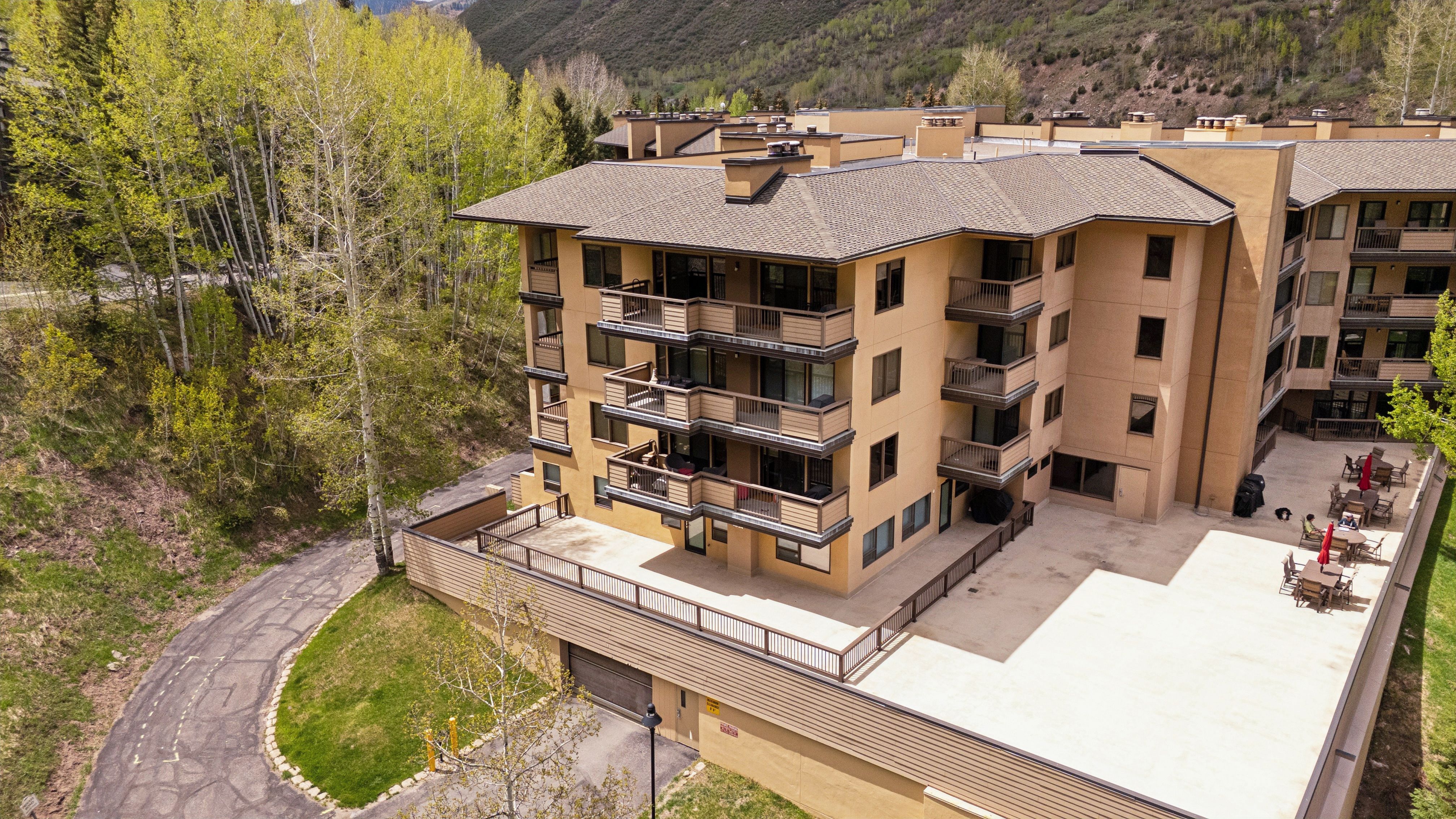 This high-angle aerial shot captures a multi-story residential complex nestled against a lush, forested hillside. The building features a tan stucco facade with tiered balconies, a large ground-level terrace, and a paved parking area. The perspective highlights the property's integration with the surrounding natural landscape, emphasizing its quiet, mountain-resort aesthetic.