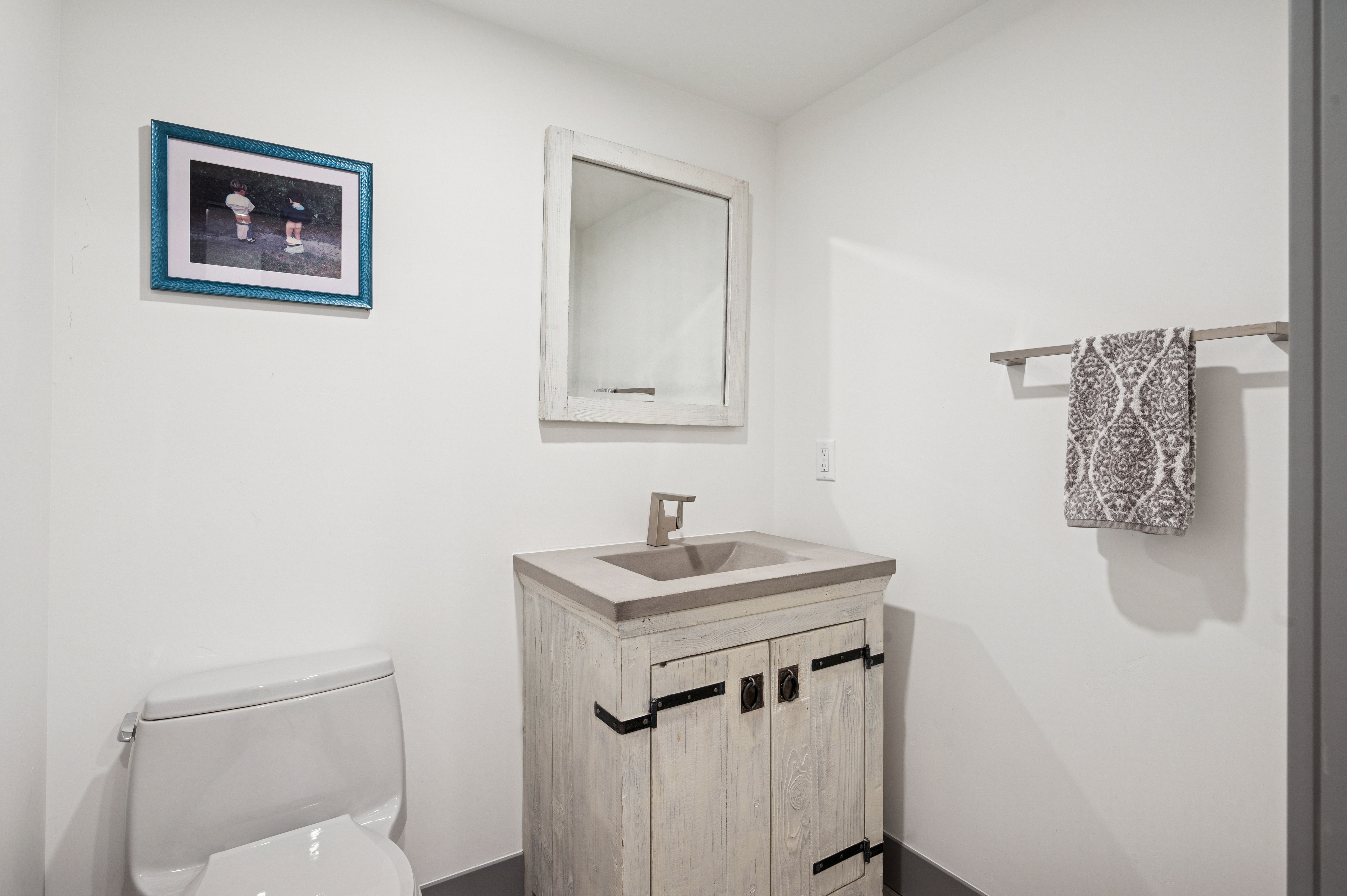 This compact powder room features a rustic-style vanity with a concrete countertop and a matching framed mirror. The space is accented by a white toilet, a decorative framed wall art piece, and a towel rack holding a patterned hand towel. The clean white walls and neutral color palette create a simple, functional, and modern farmhouse aesthetic.