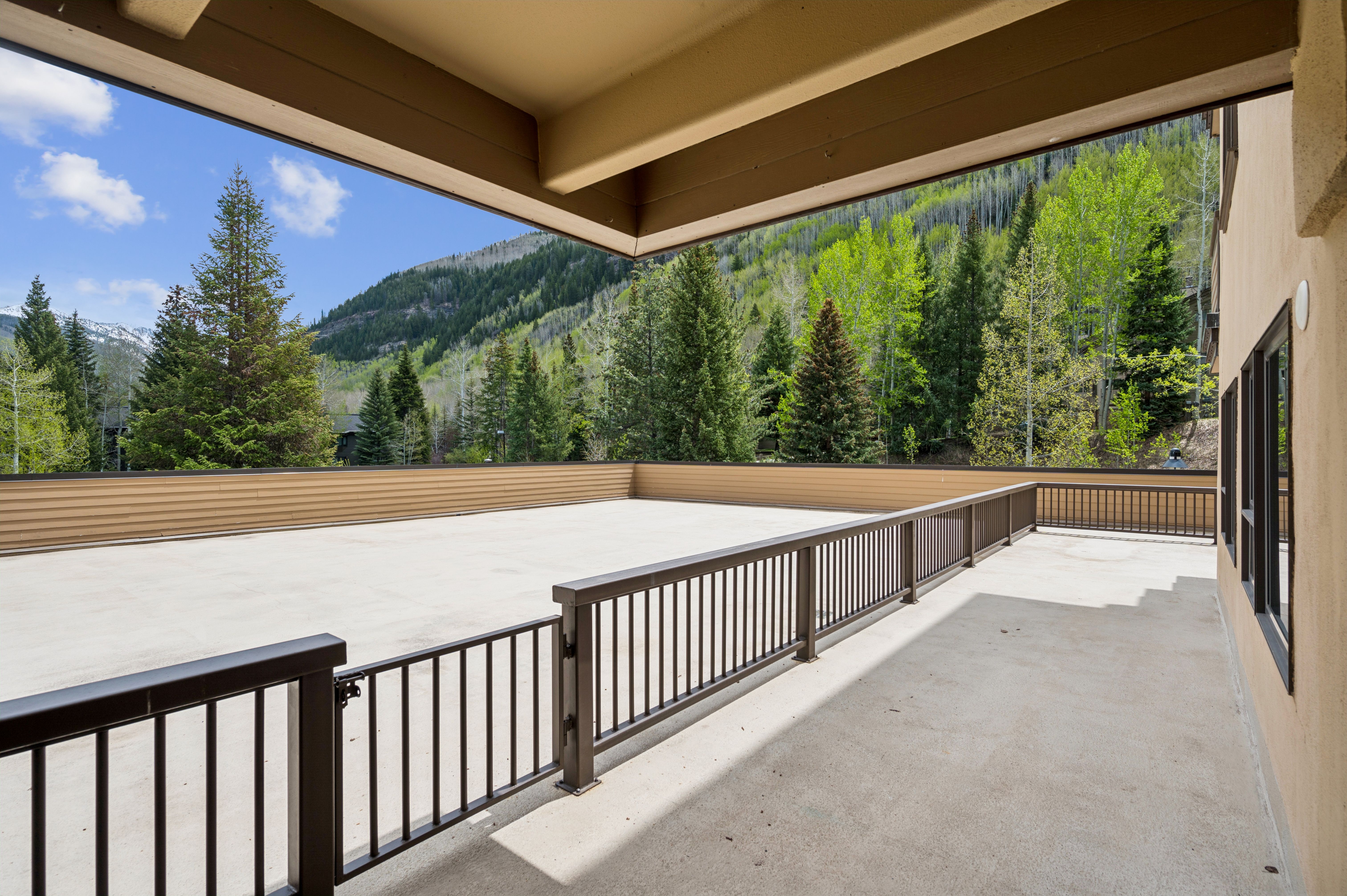 This spacious, covered balcony offers a panoramic view of a lush, mountainous landscape filled with evergreen and aspen trees. The deck features a clean, concrete surface enclosed by dark metal railings, providing a secure and open-air space for outdoor relaxation. The architectural design emphasizes a seamless connection between the home's interior and the natural beauty of the surrounding environment.