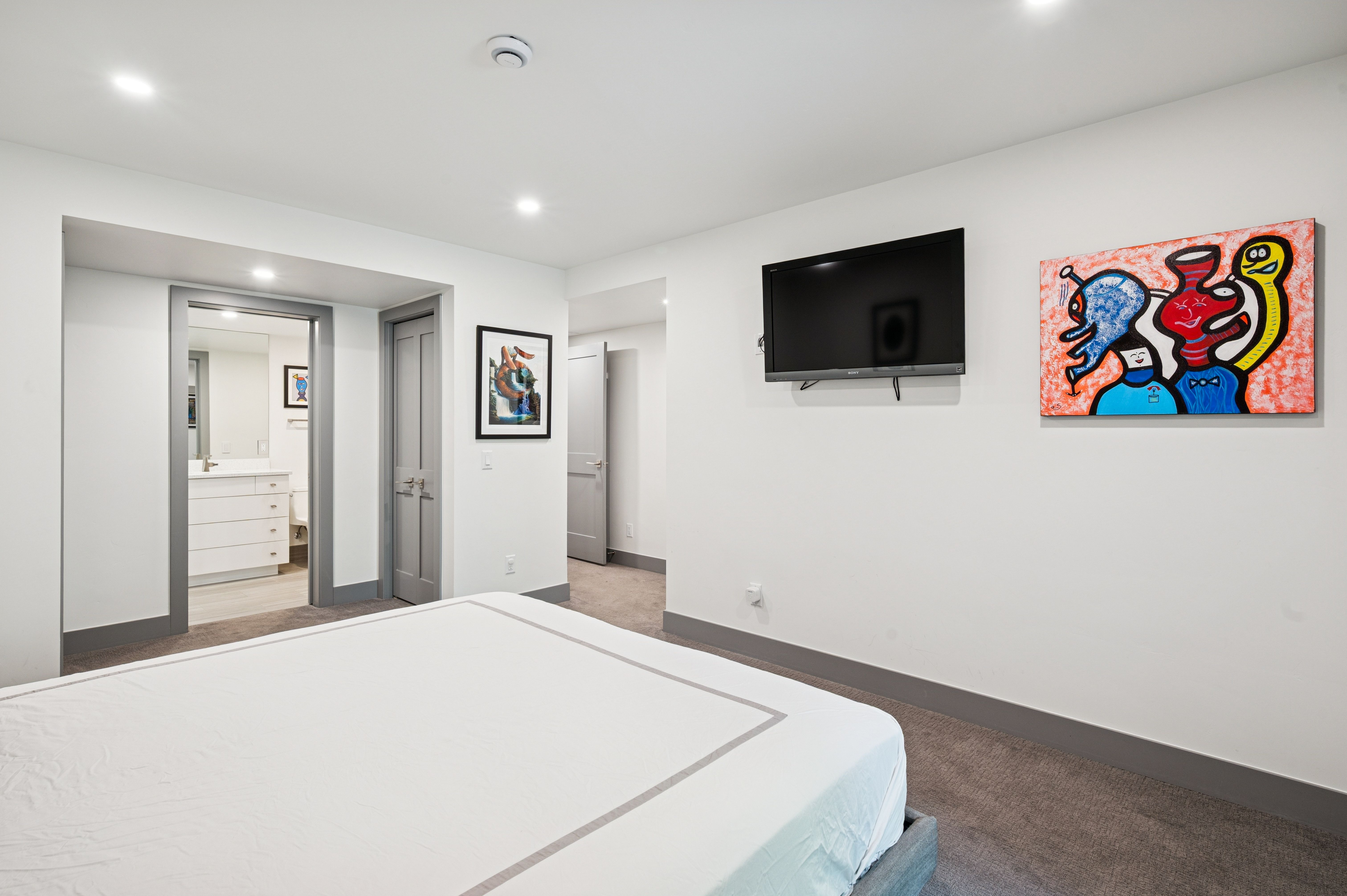 This clean and minimalist bedroom features neutral gray carpeting and white walls, creating a bright and airy atmosphere. The room is accented by vibrant, colorful abstract artwork and includes a wall-mounted television, with a doorway leading to an adjacent bathroom. The perspective is from the foot of the bed, looking toward the main wall and the transition into the en-suite space.