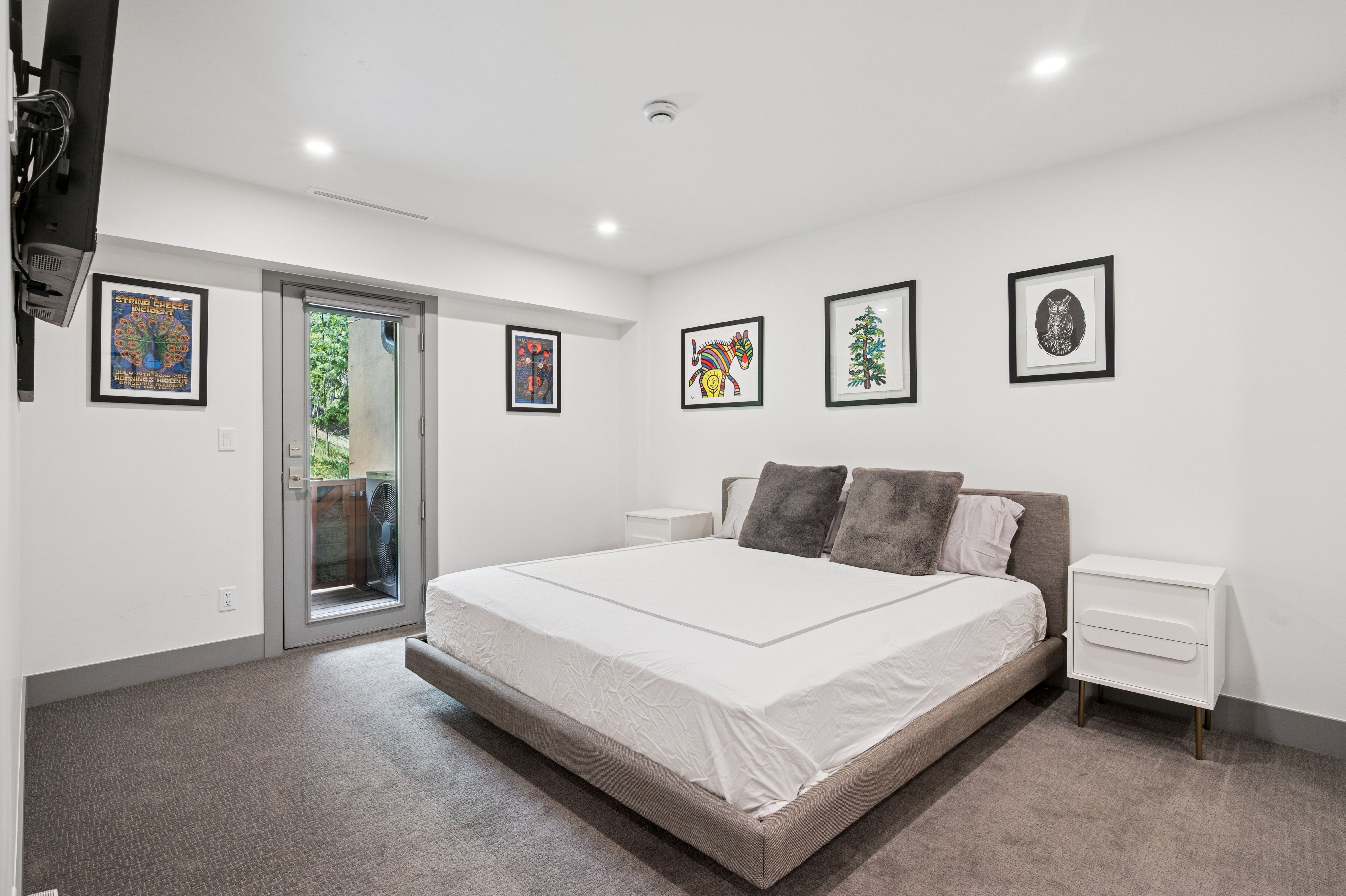This modern primary bedroom features a neutral color palette with a plush grey upholstered bed frame and matching grey carpeting. The room is accented by vibrant, framed wall art and clean white nightstands, while a glass door provides access to a private balcony. The perspective is a wide-angle shot that emphasizes the spaciousness and contemporary design of the suite.