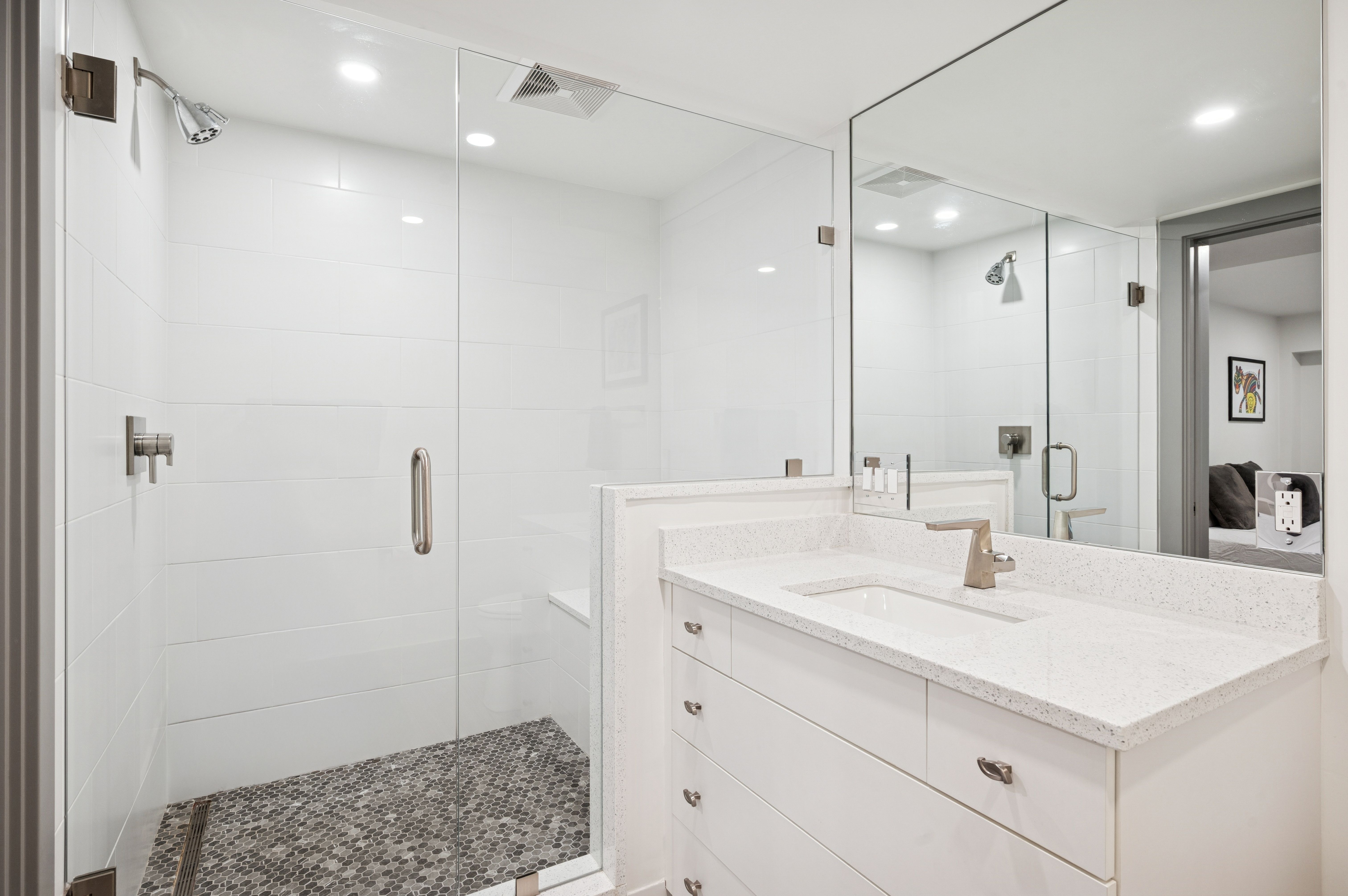 This modern, minimalist bathroom features a sleek walk-in shower with a glass enclosure and a distinctive dark pebble-tile floor. Adjacent to the shower is a clean white vanity topped with a speckled quartz countertop and a contemporary faucet. The space is bright and airy, enhanced by a large wall-to-wall mirror that reflects the clean lines and neutral color palette.