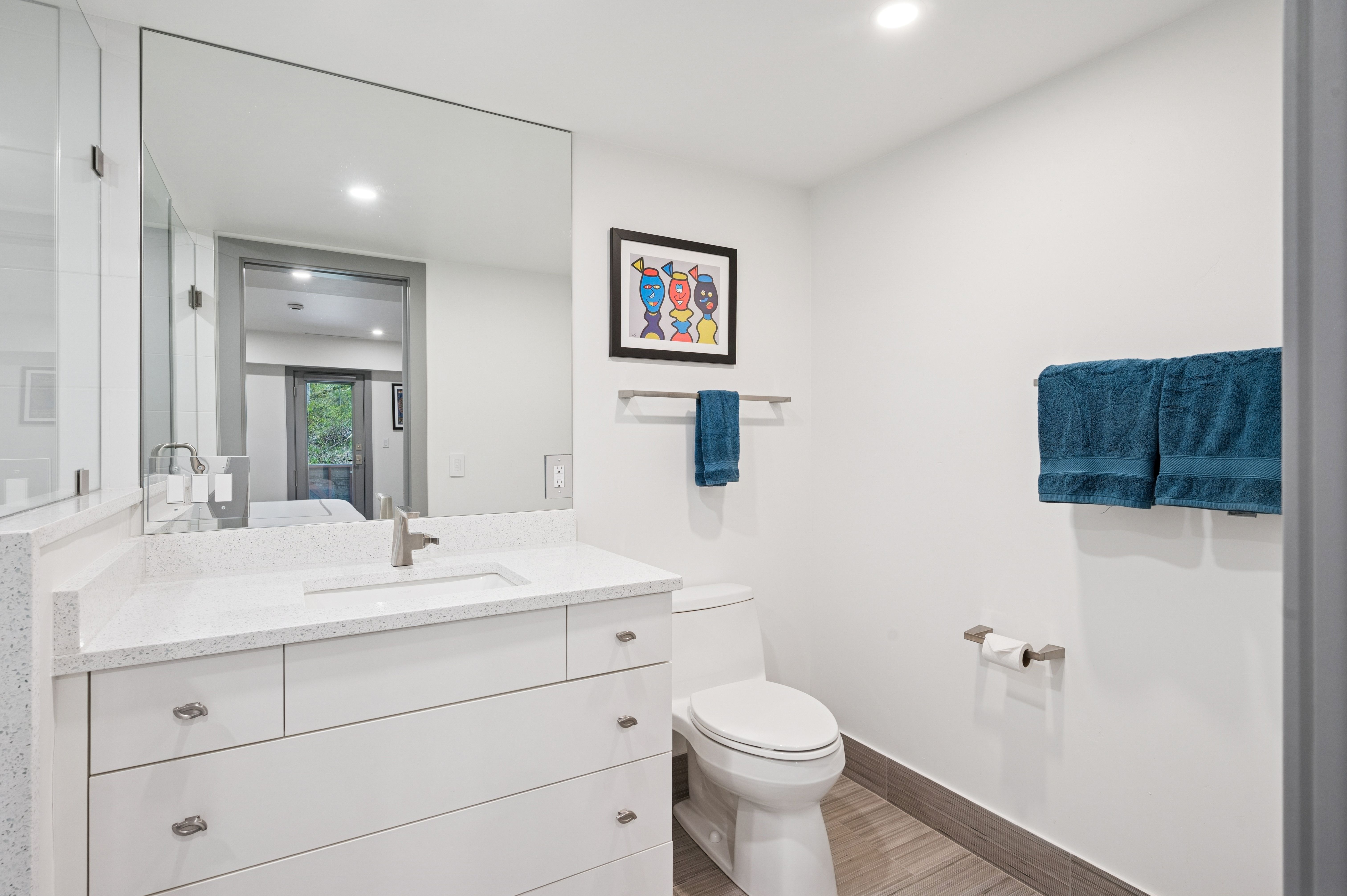 This clean and modern bathroom features a white vanity with a speckled quartz countertop, a large wall-mounted mirror, and a sleek white toilet. The space is accented with a colorful piece of framed wall art, blue towels, and wood-look tile flooring, creating a bright and functional atmosphere. The perspective is a straight-on shot that captures the entire vanity and toilet area, emphasizing the efficient layout.