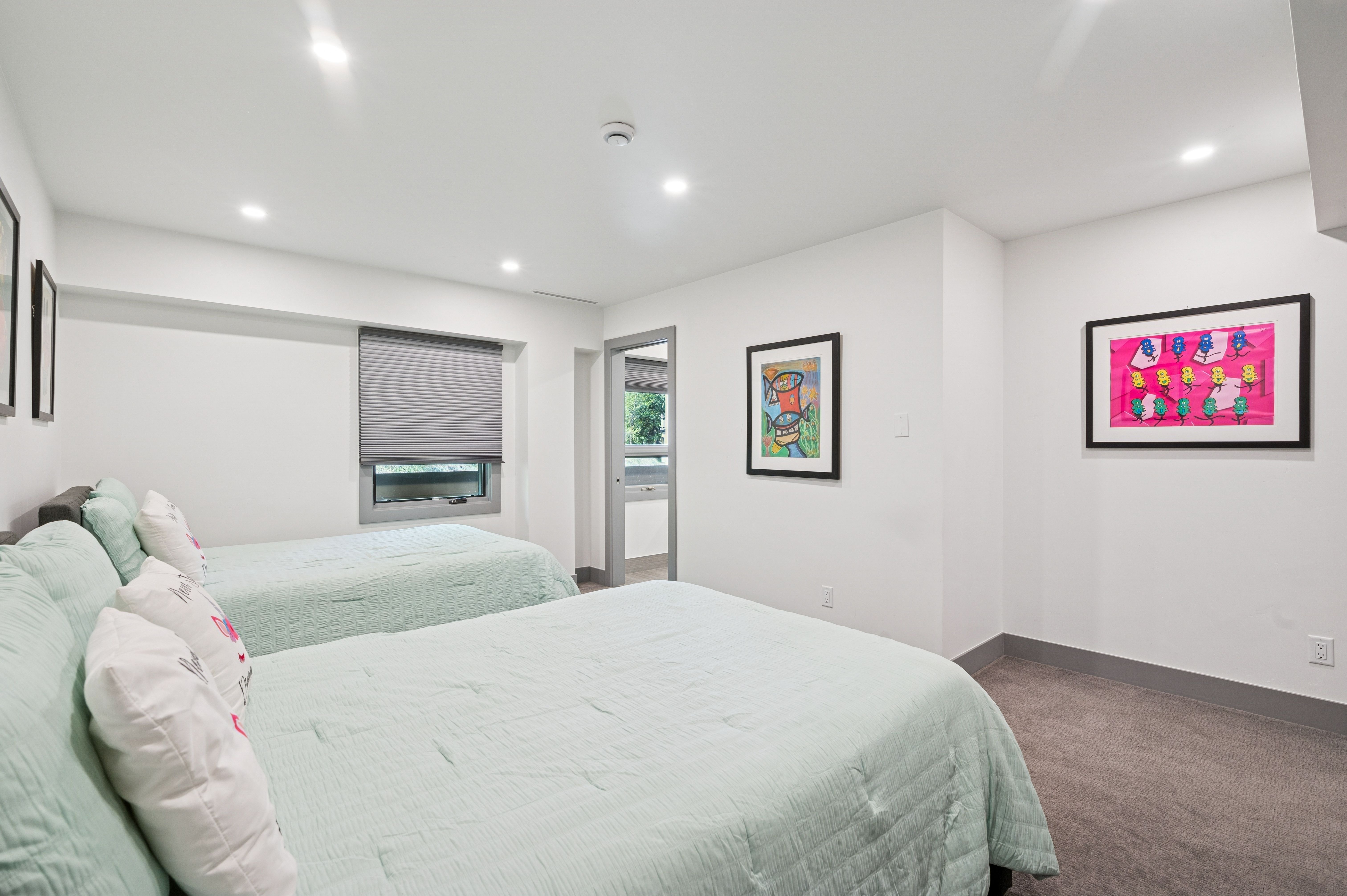 This bright and minimalist guest bedroom features two twin beds with light green quilted bedding and decorative pillows. The room is accented by colorful, modern framed artwork on the white walls and is illuminated by recessed ceiling lighting. A window with a grey pleated shade provides natural light, while the neutral carpet and clean lines create a calm, inviting atmosphere.