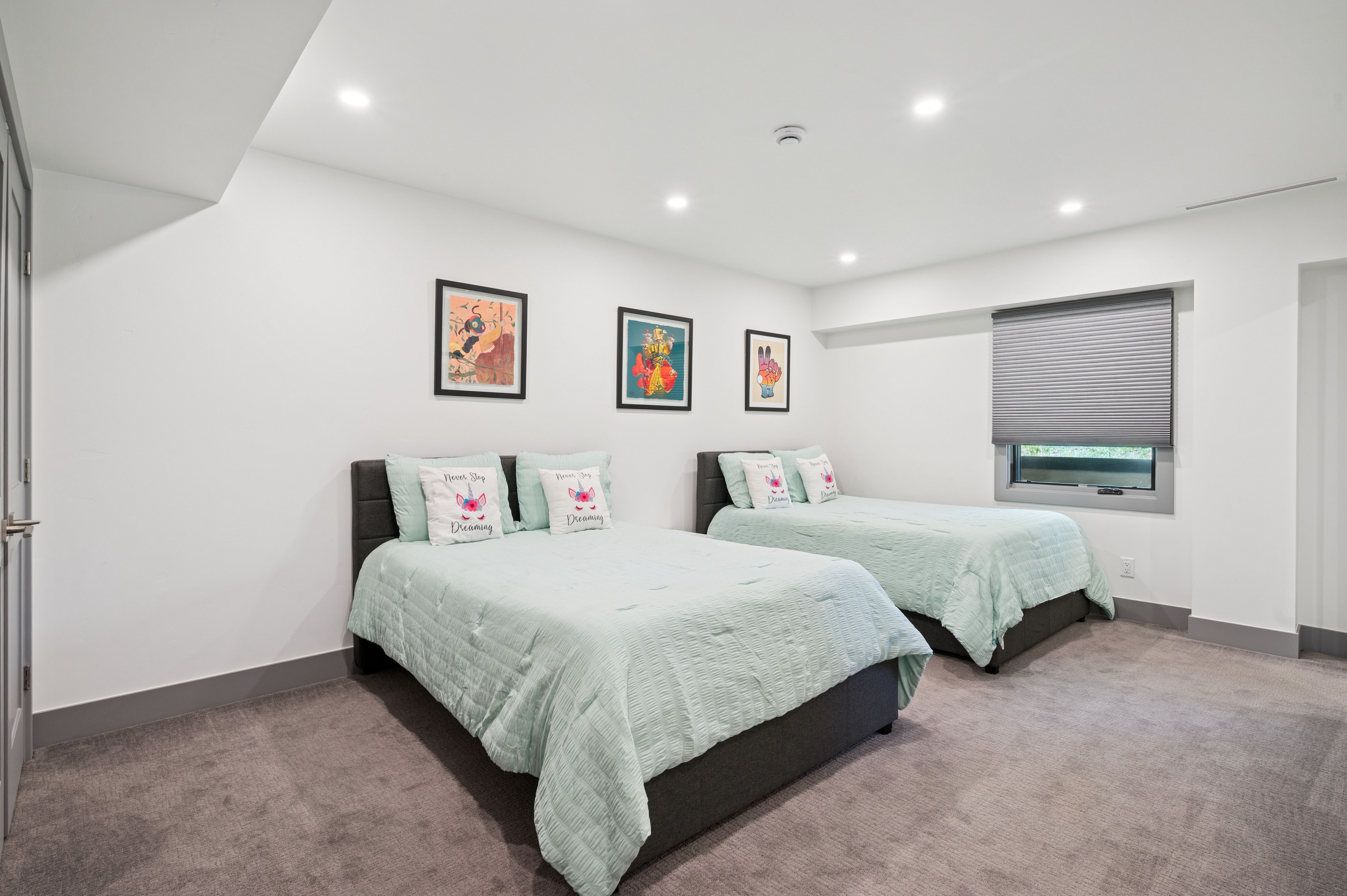 This clean and bright guest bedroom features two queen-sized beds with light mint-colored bedding and decorative unicorn-themed pillows. The room is accented by three framed art pieces on the wall, neutral grey carpeting, and a window with a modern grey shade. The minimalist design and recessed lighting create a welcoming and comfortable atmosphere.