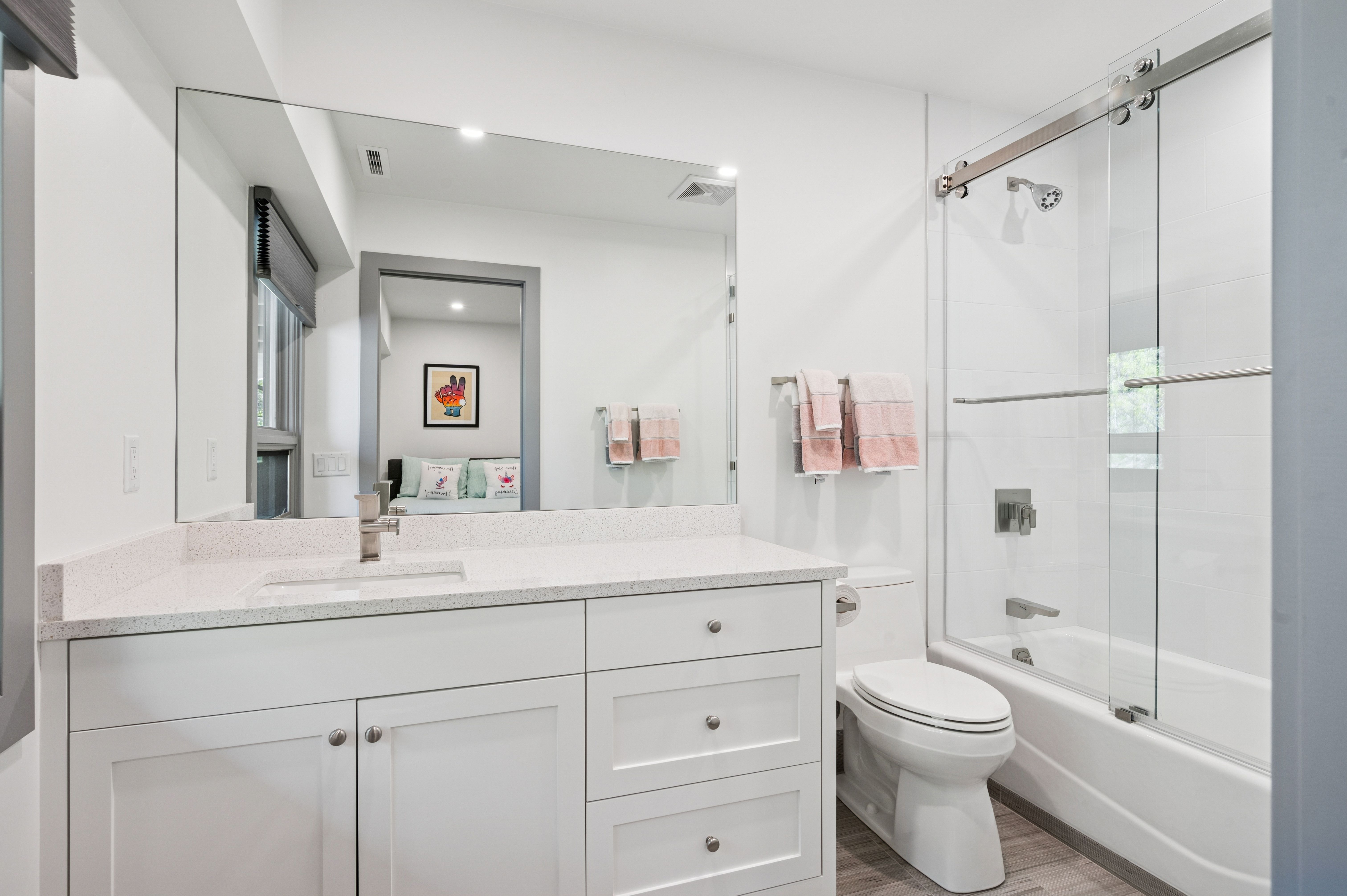 This clean and modern bathroom features a white vanity with a speckled quartz countertop, a large wall-to-wall mirror, and a shower-tub combination enclosed by a sliding glass door. The space is bright and functional, with neutral tones and contemporary fixtures that create a welcoming atmosphere. A view into the adjacent bedroom is visible through the mirror's reflection, adding depth to the composition.