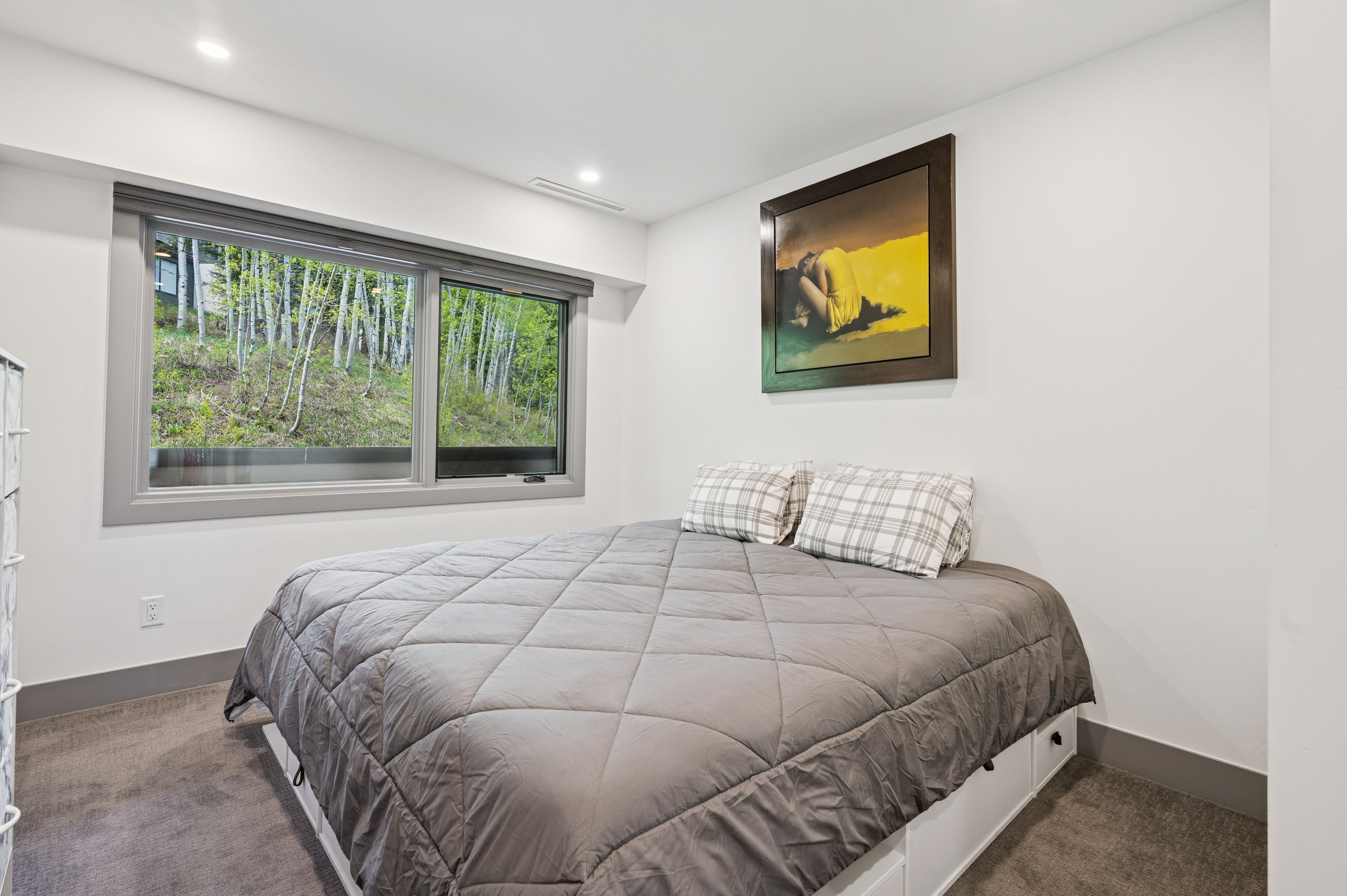This cozy bedroom features a platform bed with built-in storage drawers, dressed in a grey quilted comforter and plaid pillows. A large window provides a serene view of the surrounding forest, while a framed piece of art adds a pop of color to the minimalist white walls. The room is finished with neutral carpeting, creating a calm and inviting atmosphere.