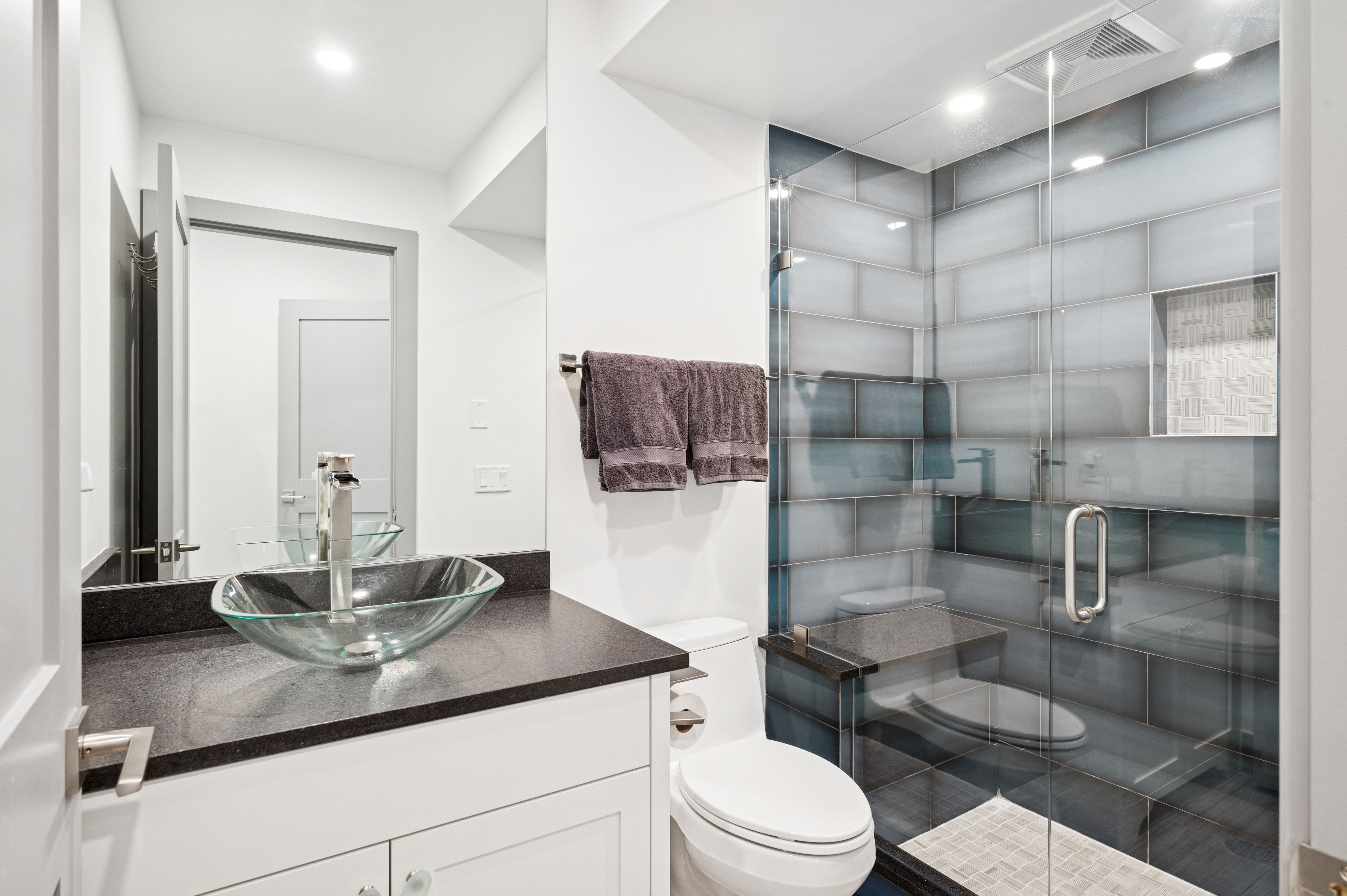 This modern bathroom features a sleek glass vessel sink atop a dark granite countertop, paired with white cabinetry for a clean, contemporary aesthetic. The walk-in shower is a standout element, showcasing large-format blue-grey subway tiles, a built-in stone bench, and a mosaic-tiled niche. The perspective captures the efficient layout, highlighting the seamless integration of the vanity, toilet, and glass-enclosed shower area.