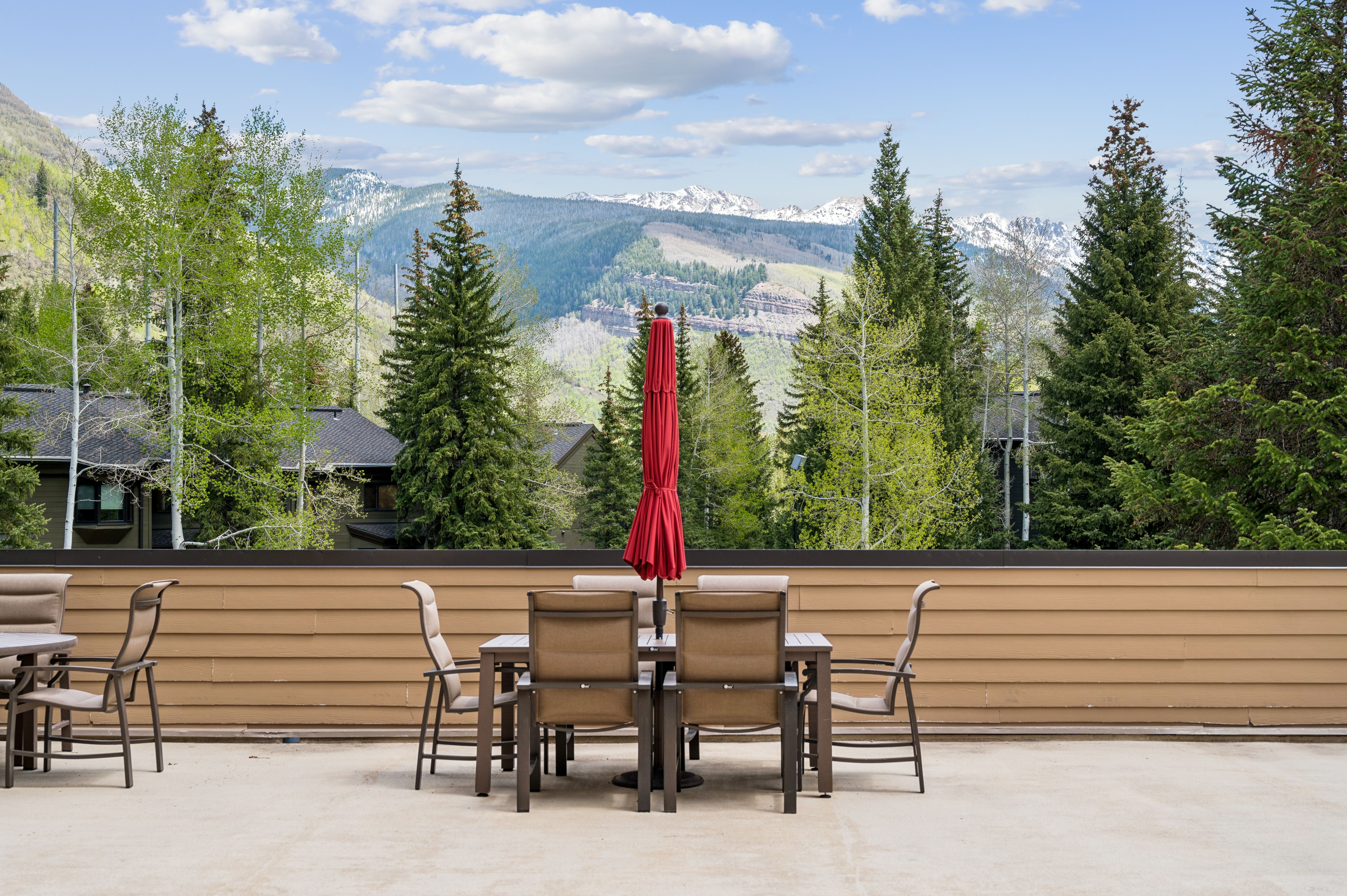 This spacious outdoor deck features a dining set with a red umbrella, perfectly positioned to take in the breathtaking panoramic views of snow-capped mountains and lush evergreen forests. The clean, neutral-toned concrete surface and simple wooden railing create a minimalist backdrop that emphasizes the natural beauty of the surrounding landscape. The perspective is eye-level, capturing a serene and inviting atmosphere ideal for outdoor dining or relaxation.