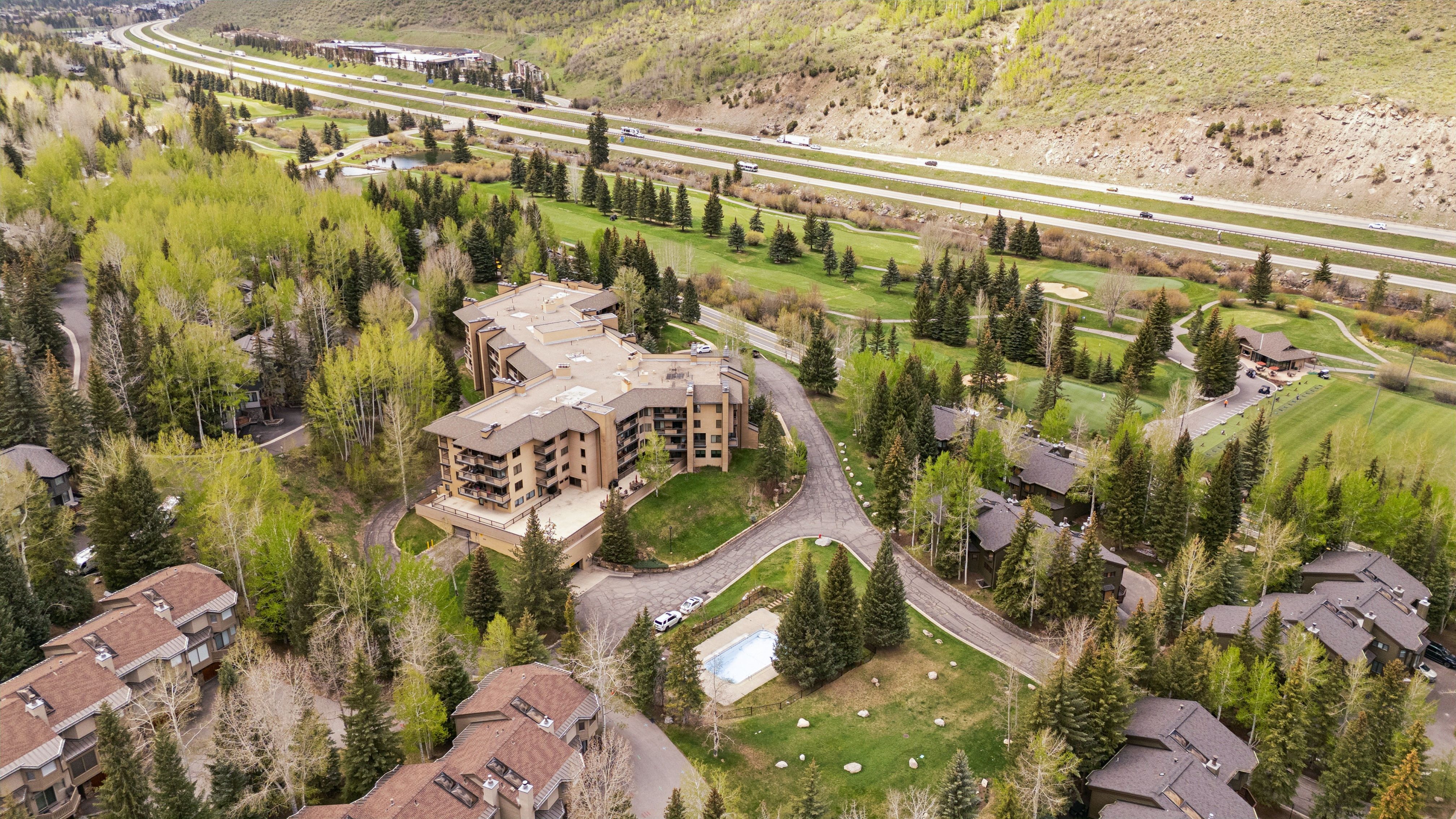 This high-angle aerial view captures a multi-story residential complex nestled within a lush, mountainous landscape. The property features a central building with balconies, a private swimming pool, and winding paved driveways, all surrounded by dense pine forests and a nearby golf course. The scene is framed by a major highway in the distance, emphasizing the property's secluded yet accessible location in a scenic mountain setting.