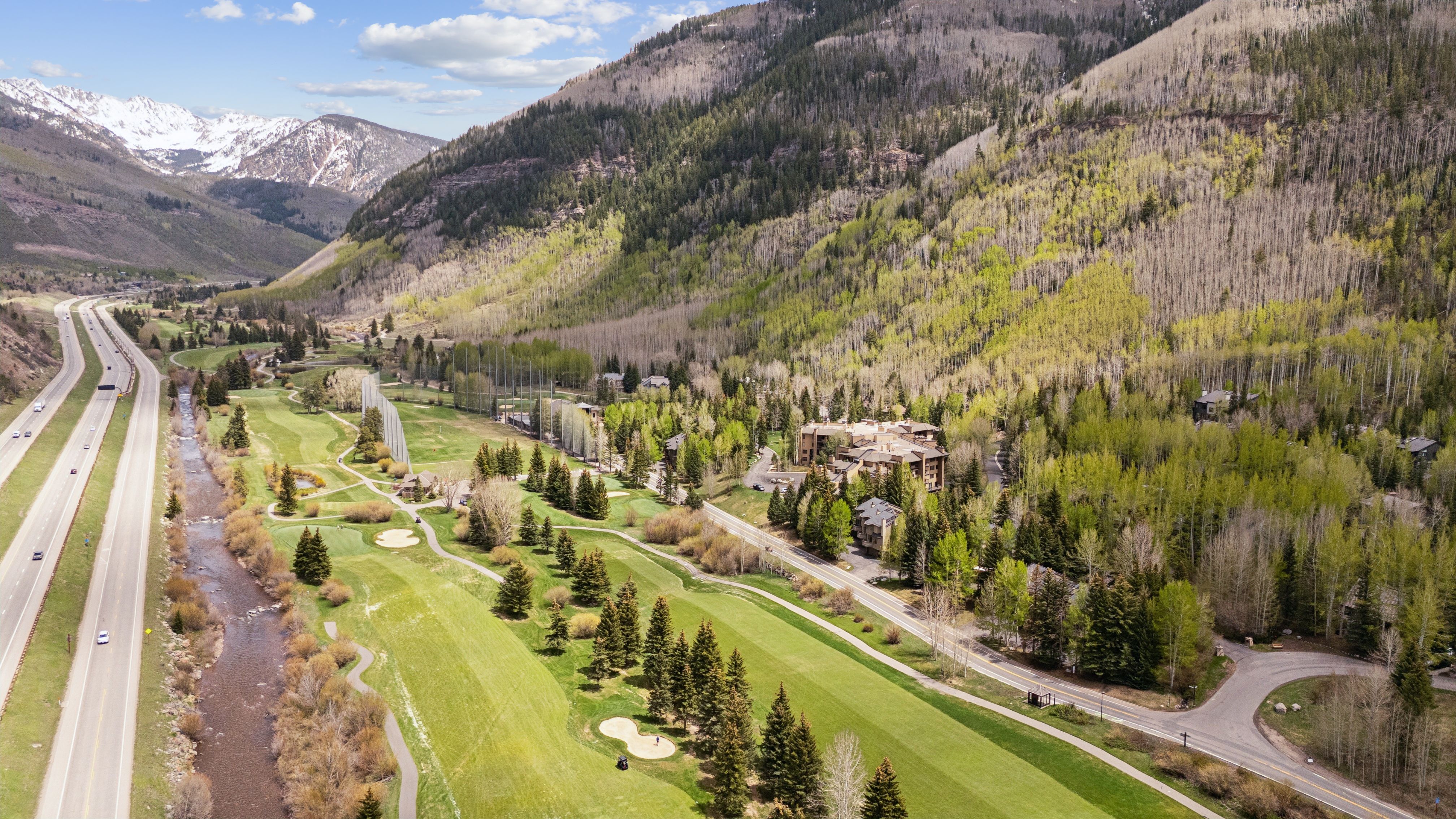 This high-angle aerial shot captures a scenic mountain valley featuring a lush golf course nestled between a winding river and a multi-lane highway. The landscape is characterized by dense evergreen and deciduous forests, with snow-capped peaks looming in the distance. The perspective emphasizes the expansive, tranquil setting of the property, highlighting its integration with the natural environment.