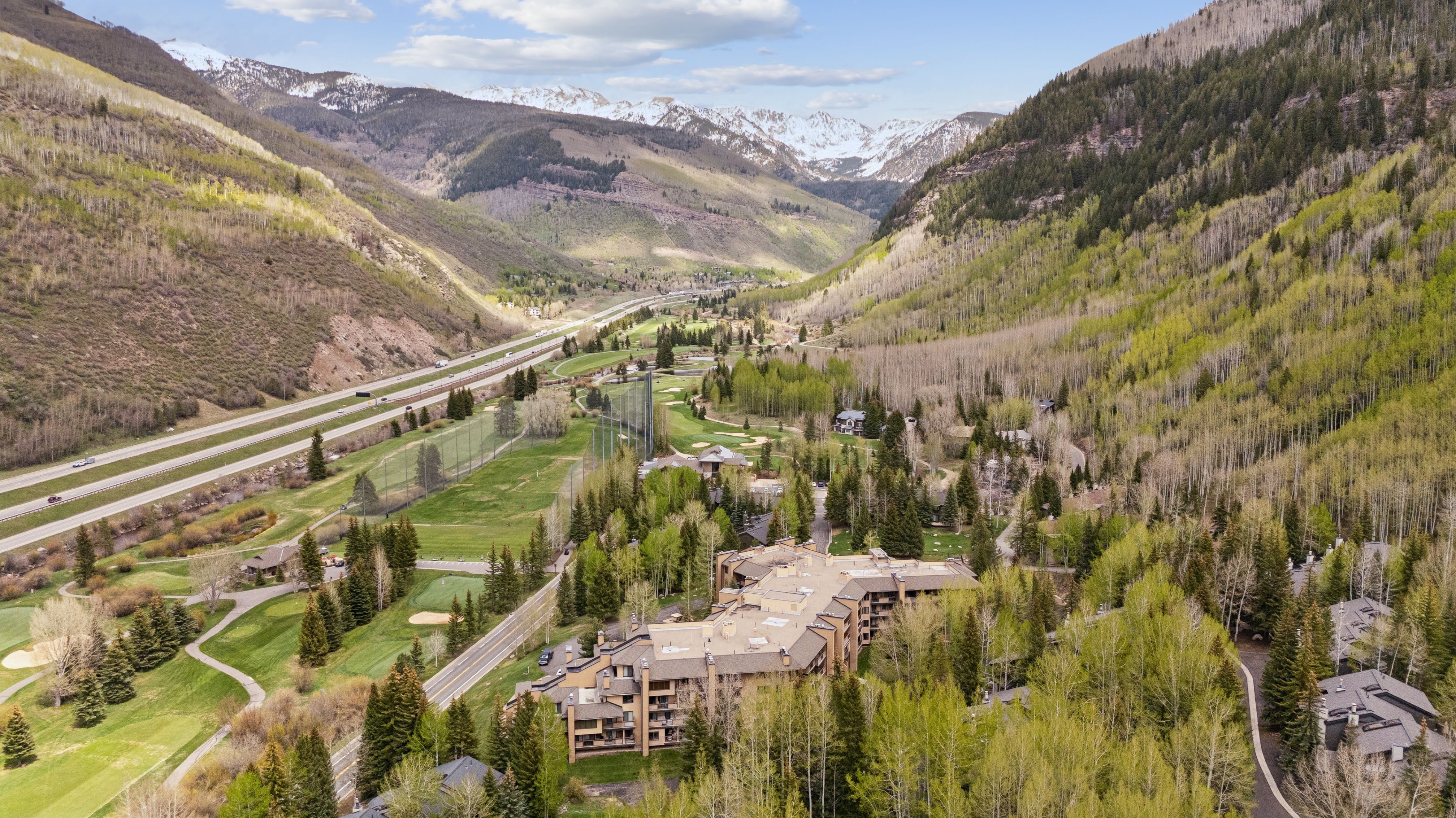 This high-angle aerial shot captures a sprawling mountain resort complex nestled within a lush, green valley surrounded by towering, snow-capped peaks. The scene features a multi-story lodge with a classic alpine architectural style, situated adjacent to a golf course and a major highway that winds through the landscape. The combination of dense forest, expansive mountain vistas, and well-maintained recreational grounds creates a serene and inviting atmosphere for a mountain getaway.
