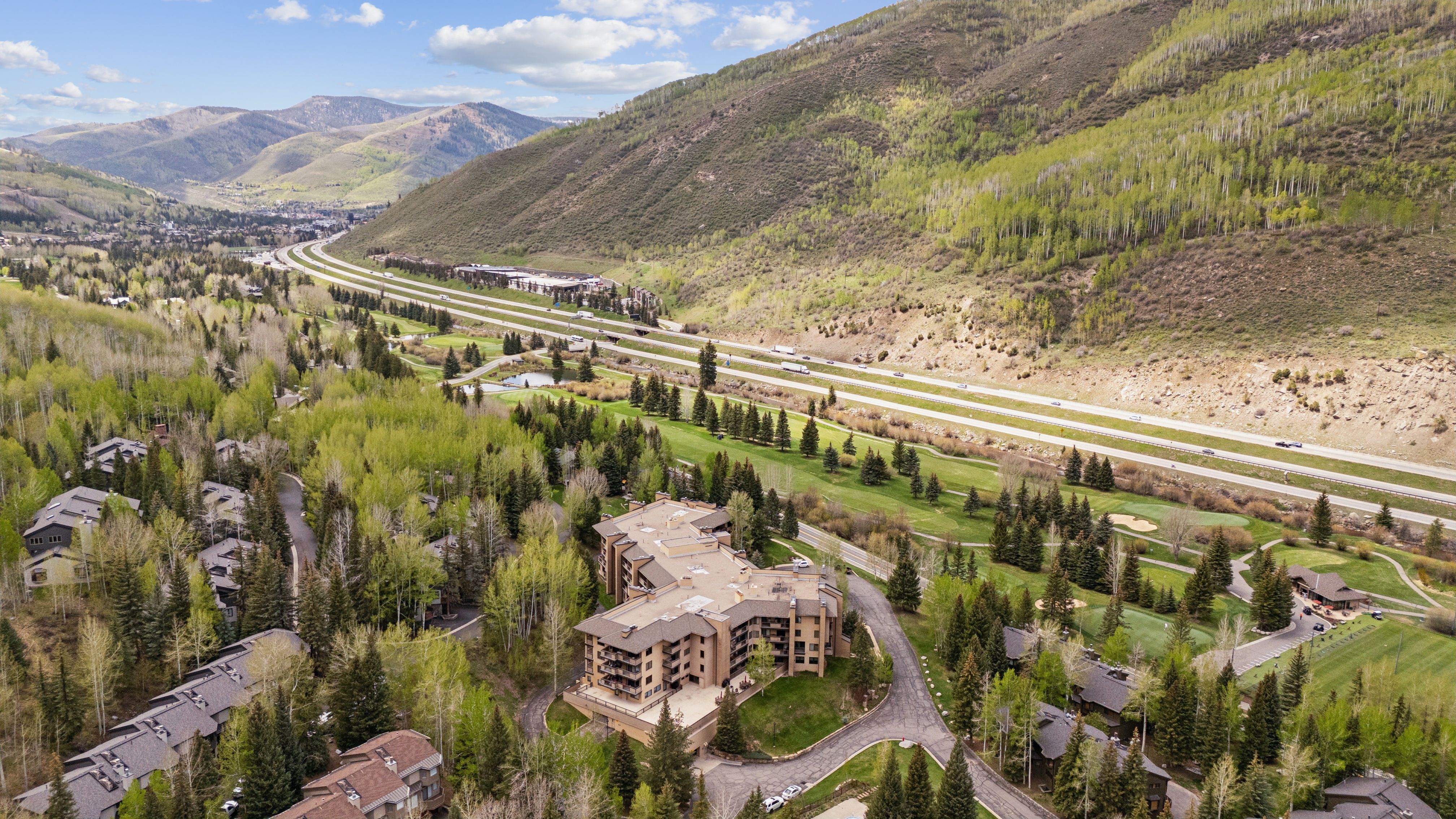 This high-angle aerial shot captures a multi-story residential complex nestled within a lush, forested mountain valley. The property is situated adjacent to a golf course and a major highway, offering a blend of natural serenity and convenient accessibility. The surrounding landscape features dense evergreen and aspen trees, with rolling mountains in the distance under a bright, partly cloudy sky.