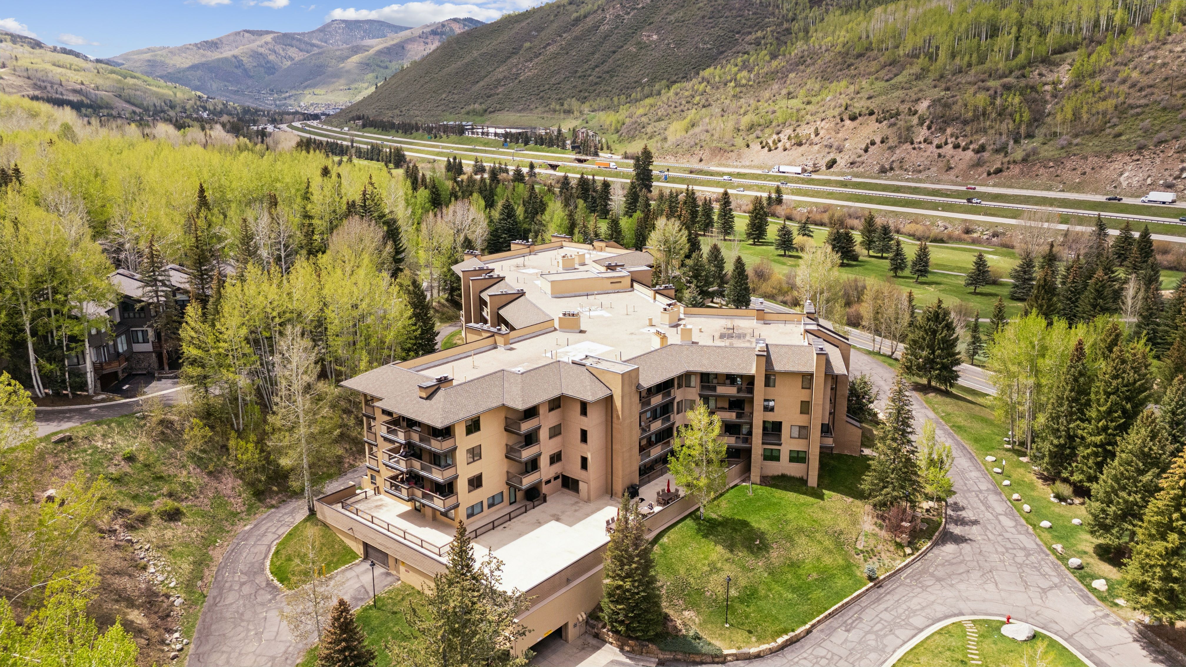 This high-angle aerial shot captures a multi-story residential complex nestled in a lush, mountainous landscape. The building features a tan facade with numerous balconies and a flat roof, surrounded by dense evergreen and aspen trees. In the background, a highway winds through the valley, emphasizing the property's scenic and accessible location.