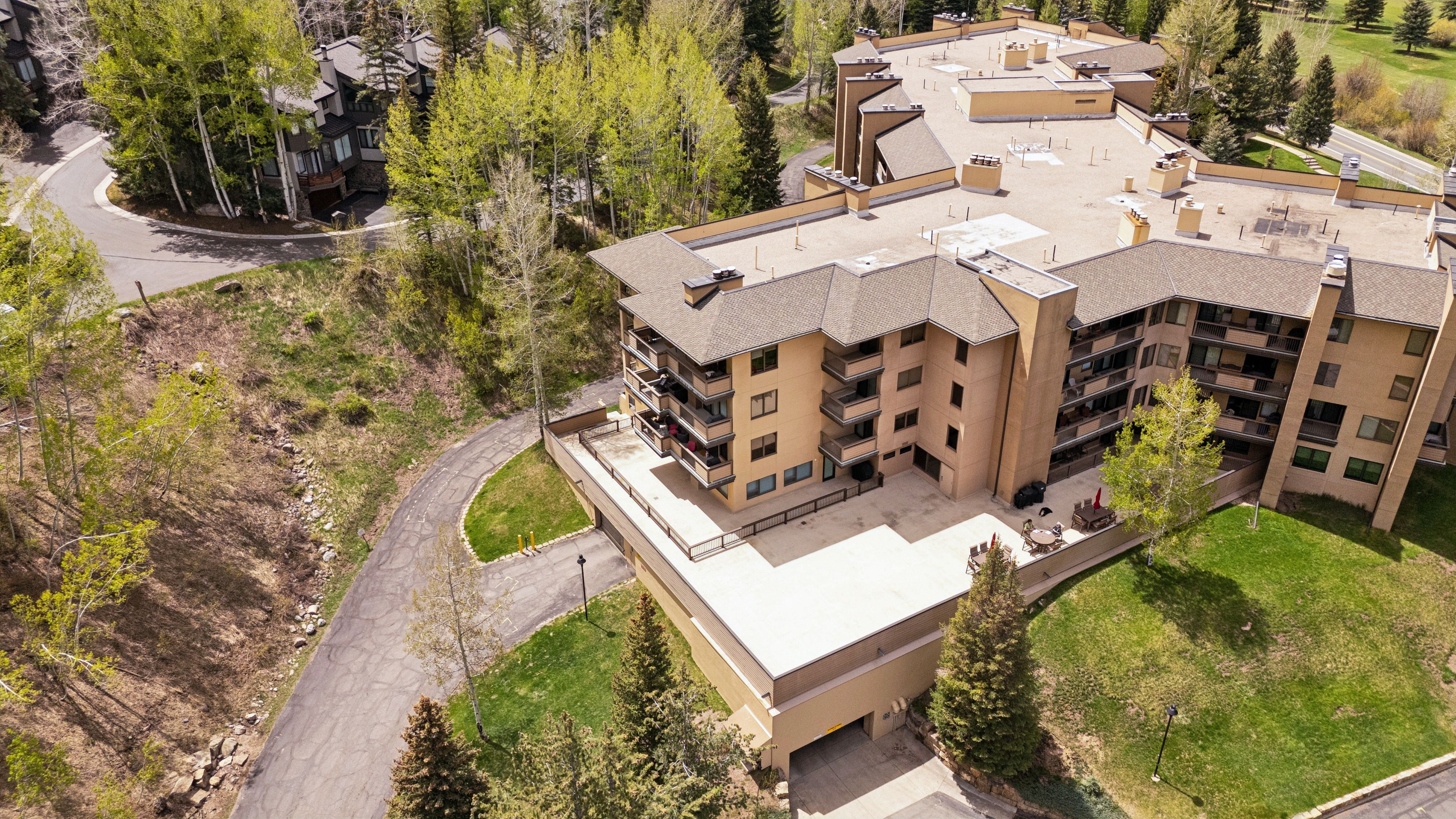 This high-angle aerial view captures a multi-story residential complex nestled in a lush, wooded landscape. The building features a tan stucco facade with multiple balconies, a large flat rooftop, and an expansive ground-level patio area. The surrounding grounds include paved driveways, manicured lawns, and mature evergreen and deciduous trees, suggesting a serene, mountain-resort setting.