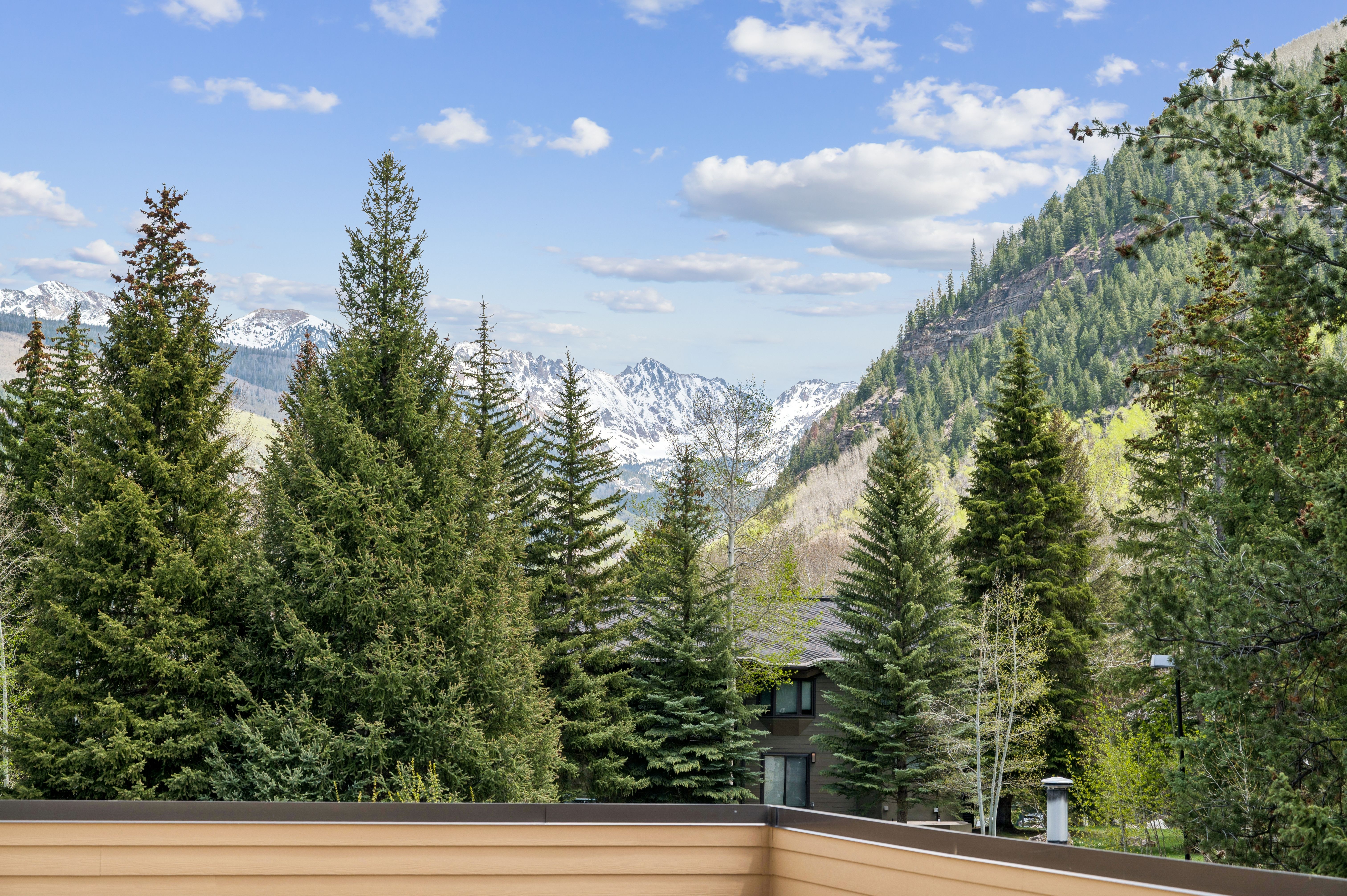 This image is captured from an elevated balcony, offering a breathtaking, panoramic view of snow-capped mountain peaks framed by lush evergreen trees. In the foreground, a clean, tan-colored railing provides a sense of perspective, while a multi-story building is partially visible nestled among the dense forest below. The scene evokes a serene, high-altitude mountain retreat atmosphere, perfect for showcasing a property's scenic location.