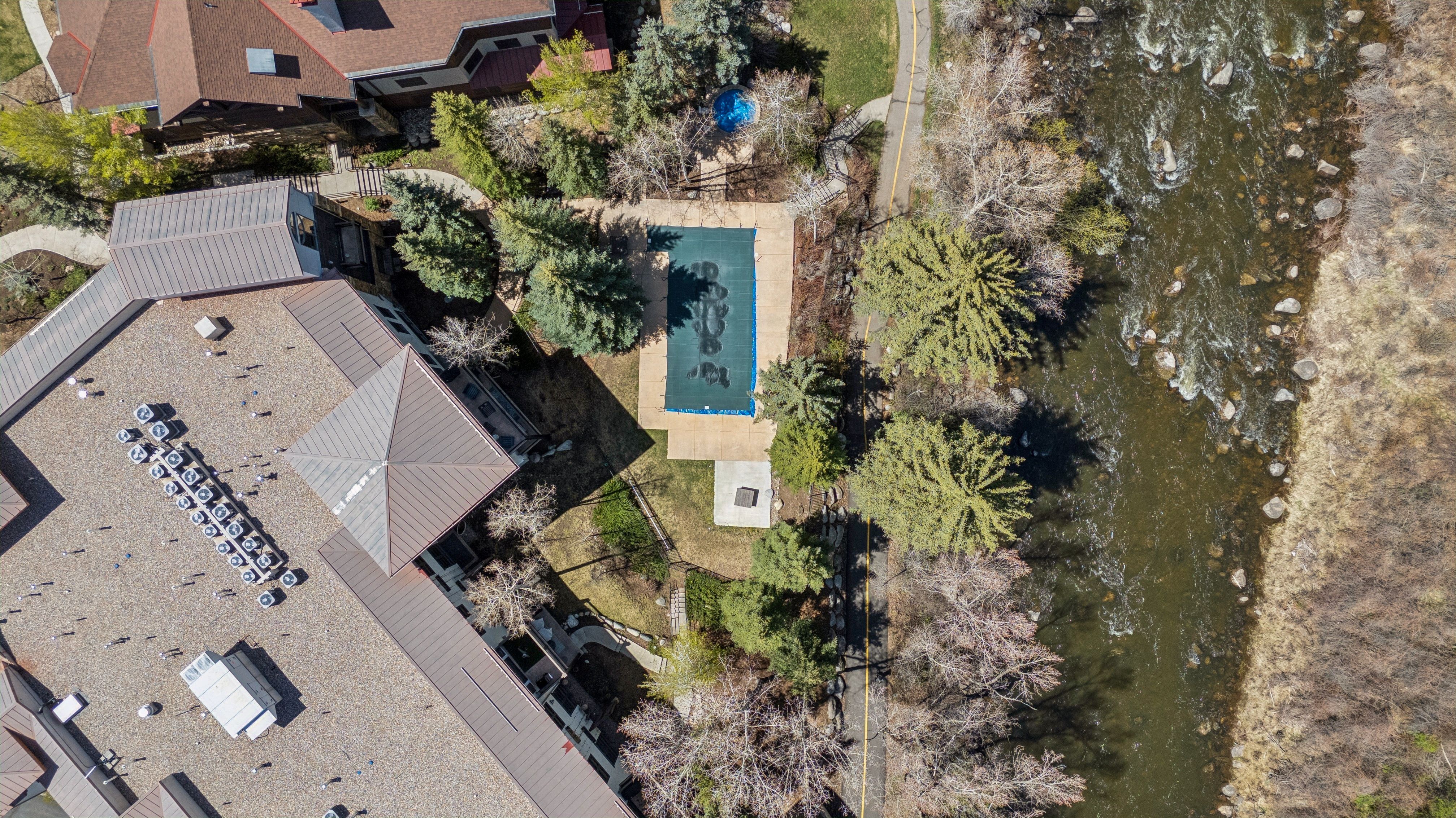 This aerial shot showcases a property featuring a large building with a complex roof structure, including multiple air conditioning units. A covered pool is nestled among mature trees, adjacent to a river or stream. A walking path runs alongside the water, enhancing the property's appeal.