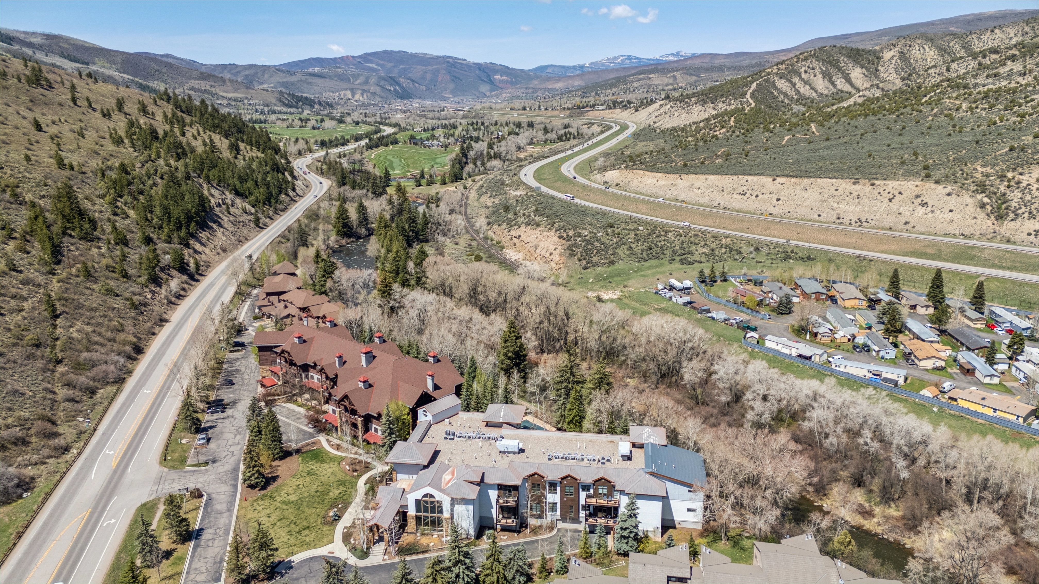 This aerial shot showcases a multi-building property nestled in a valley, featuring a mix of residential and resort-style architecture. The buildings have a combination of brown and gray roofs, complemented by lush greenery and a winding road. The surrounding landscape includes mountains and a golf course, creating a scenic and desirable location.