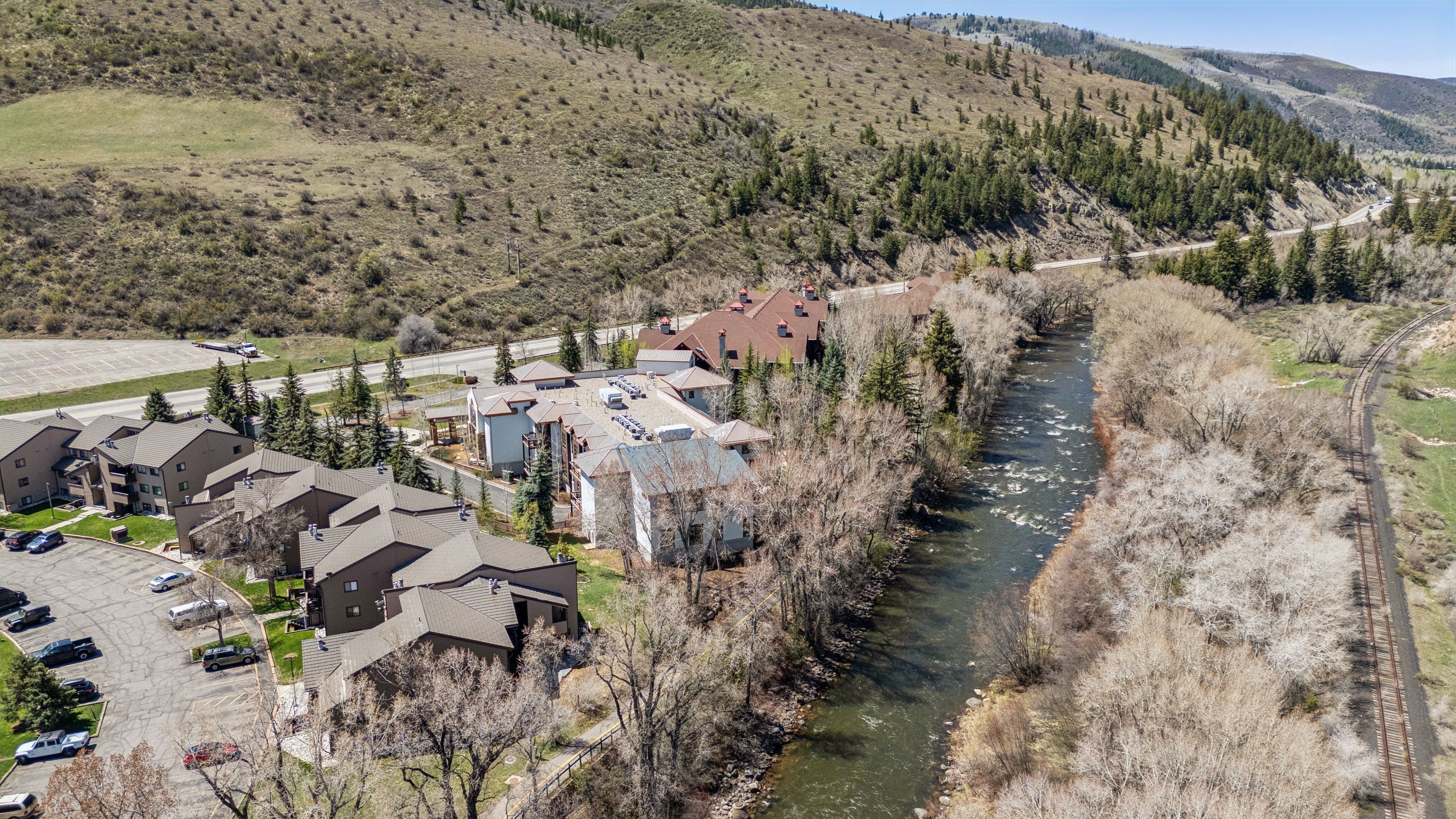 This aerial shot showcases a residential complex nestled beside a river and railroad tracks, with a backdrop of rolling hills and scattered trees. The buildings feature a mix of architectural styles, including multi-story apartments and individual homes, complemented by ample parking and landscaping. The scene conveys a sense of serene living in a natural setting.