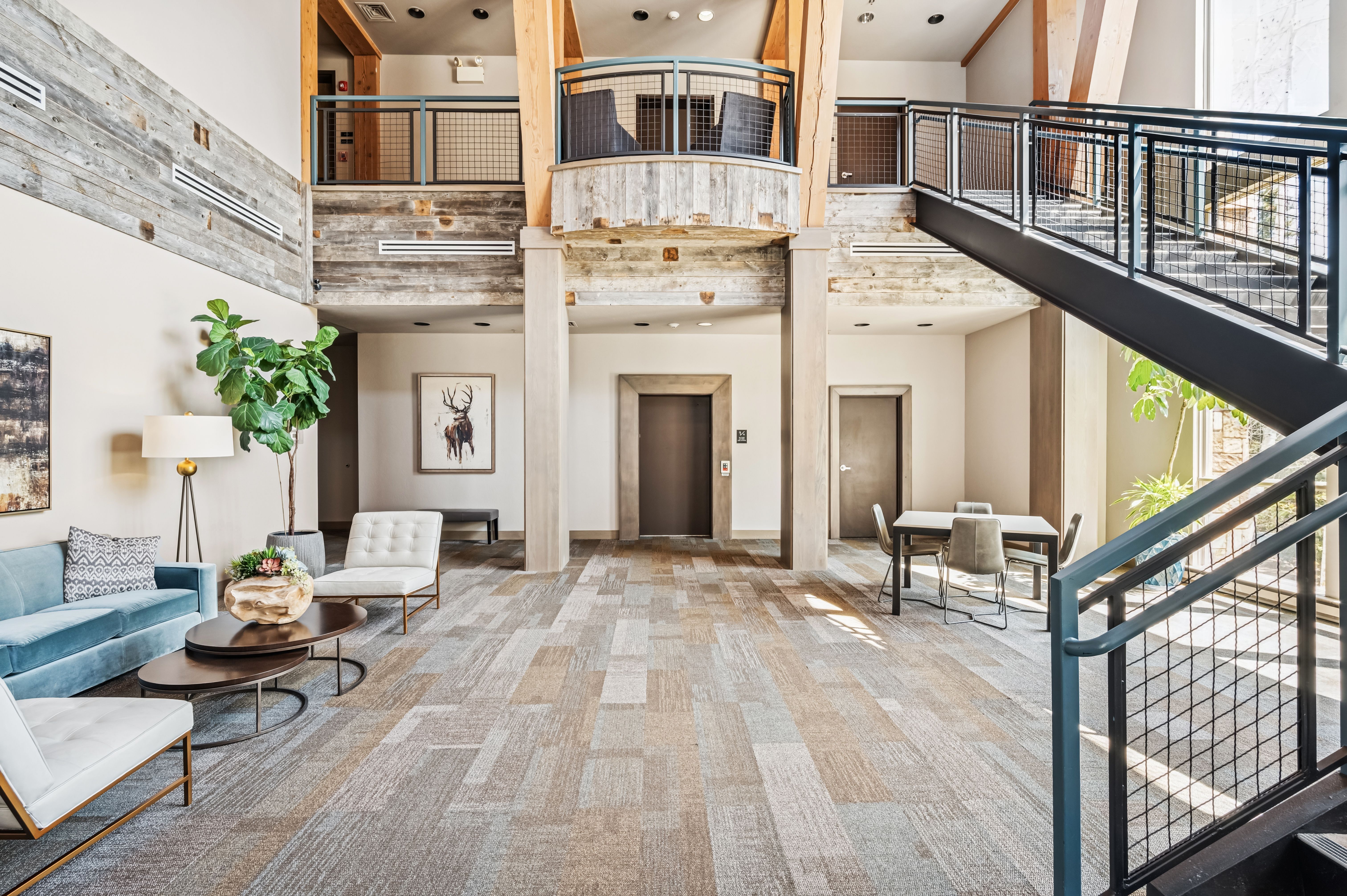 This is an interior shot of a spacious lobby or hallway area, featuring a blend of modern and rustic design elements. The area includes a seating area with a blue sofa and white chairs, a staircase with metal railings, and wooden accent walls. The flooring is a patterned carpet, and there are elevator doors and doorways visible, suggesting a multi-story building.