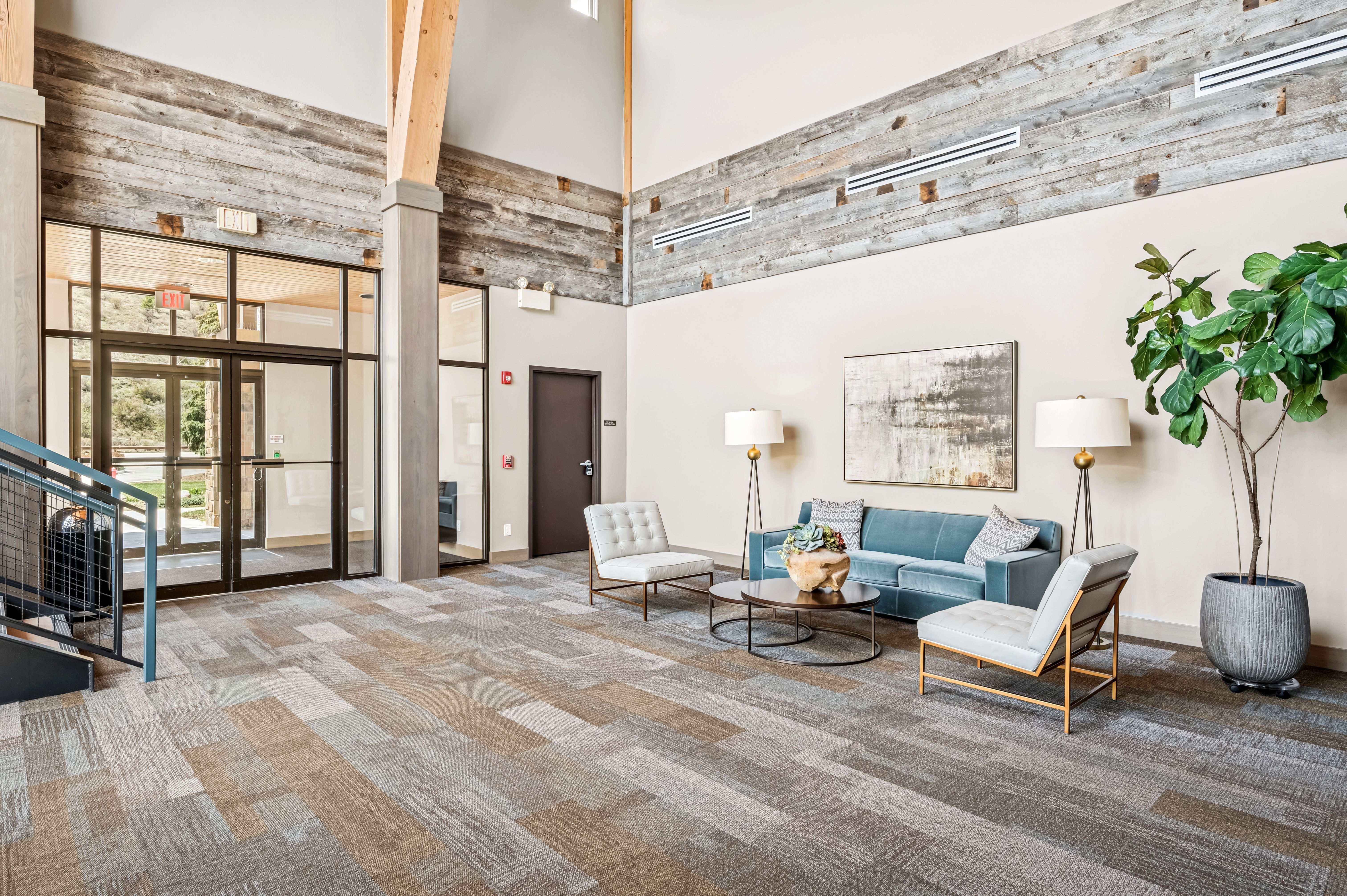 This is a spacious and modern living room area, featuring a blue velvet sofa, two white armchairs with gold frames, and a round coffee table. The room is well-lit with two floor lamps and natural light, complemented by a large abstract art piece on the wall and a tall potted plant. The flooring is a patterned carpet, and the room has a high ceiling with wooden beam accents, creating a welcoming and stylish atmosphere.