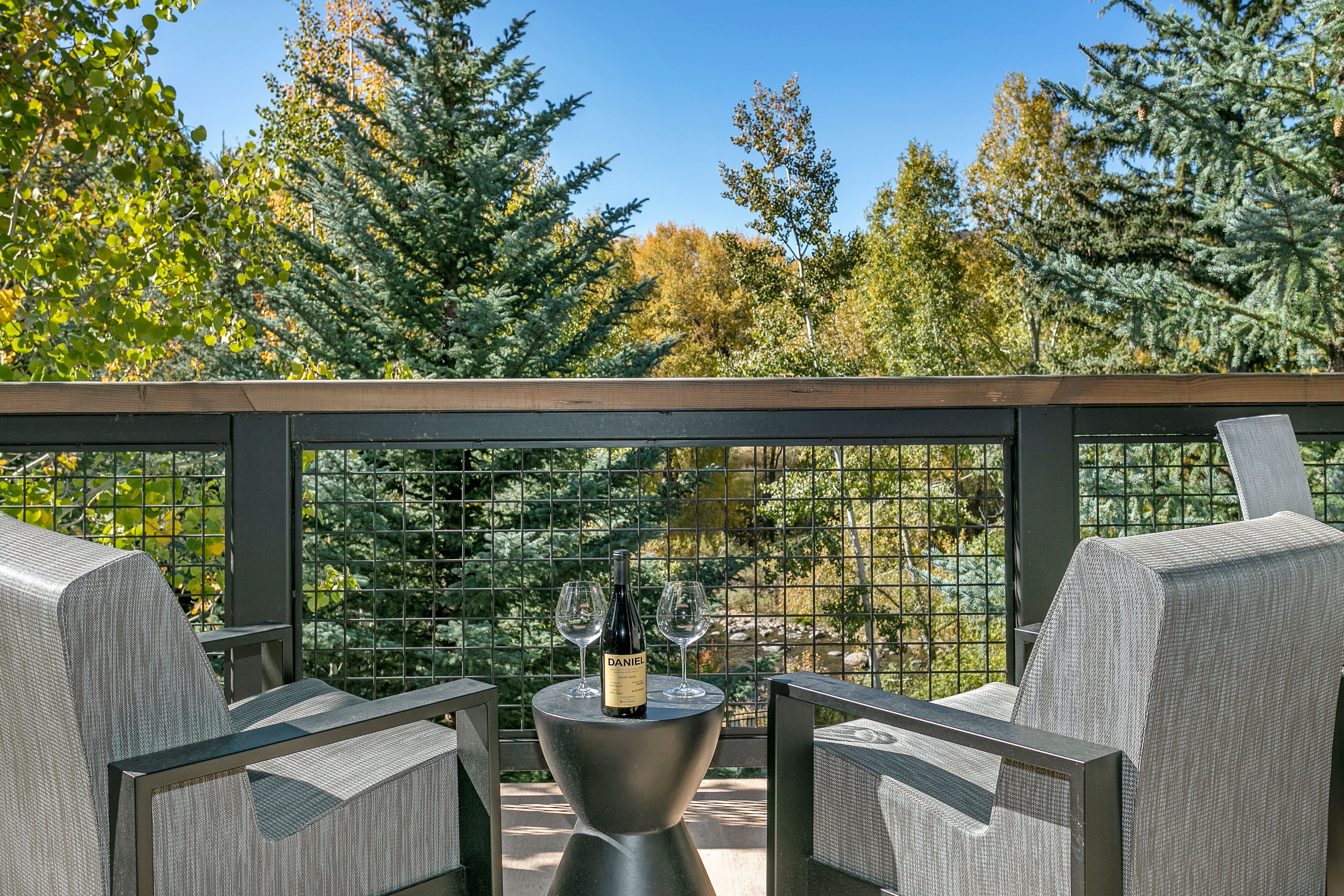 This image showcases a serene outdoor patio or balcony setting, perfect for relaxation. Two modern gray chairs flank a small, stylish black table holding a wine bottle and two glasses, suggesting an inviting space for enjoying a drink. The backdrop features a metal railing and lush greenery, creating a sense of privacy and connection with nature.