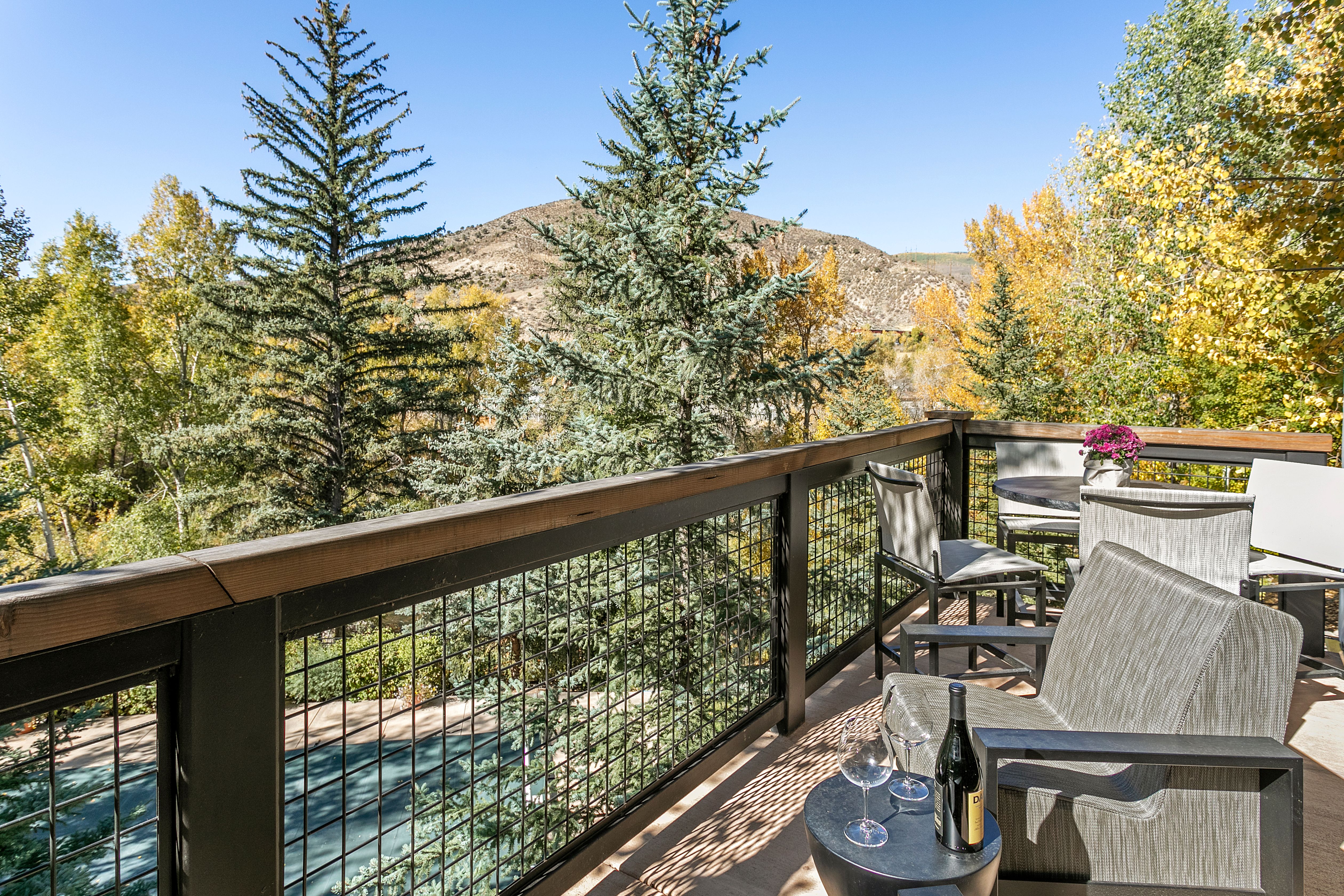 This image showcases a charming outdoor balcony or deck area, furnished with comfortable seating and a small table set with wine glasses and a bottle, suggesting relaxation and enjoyment of the surrounding natural beauty. The railing, constructed with a wood top and metal mesh, offers both safety and an unobstructed view of the trees and distant mountain. The overall impression is one of tranquility and a perfect spot for outdoor leisure.