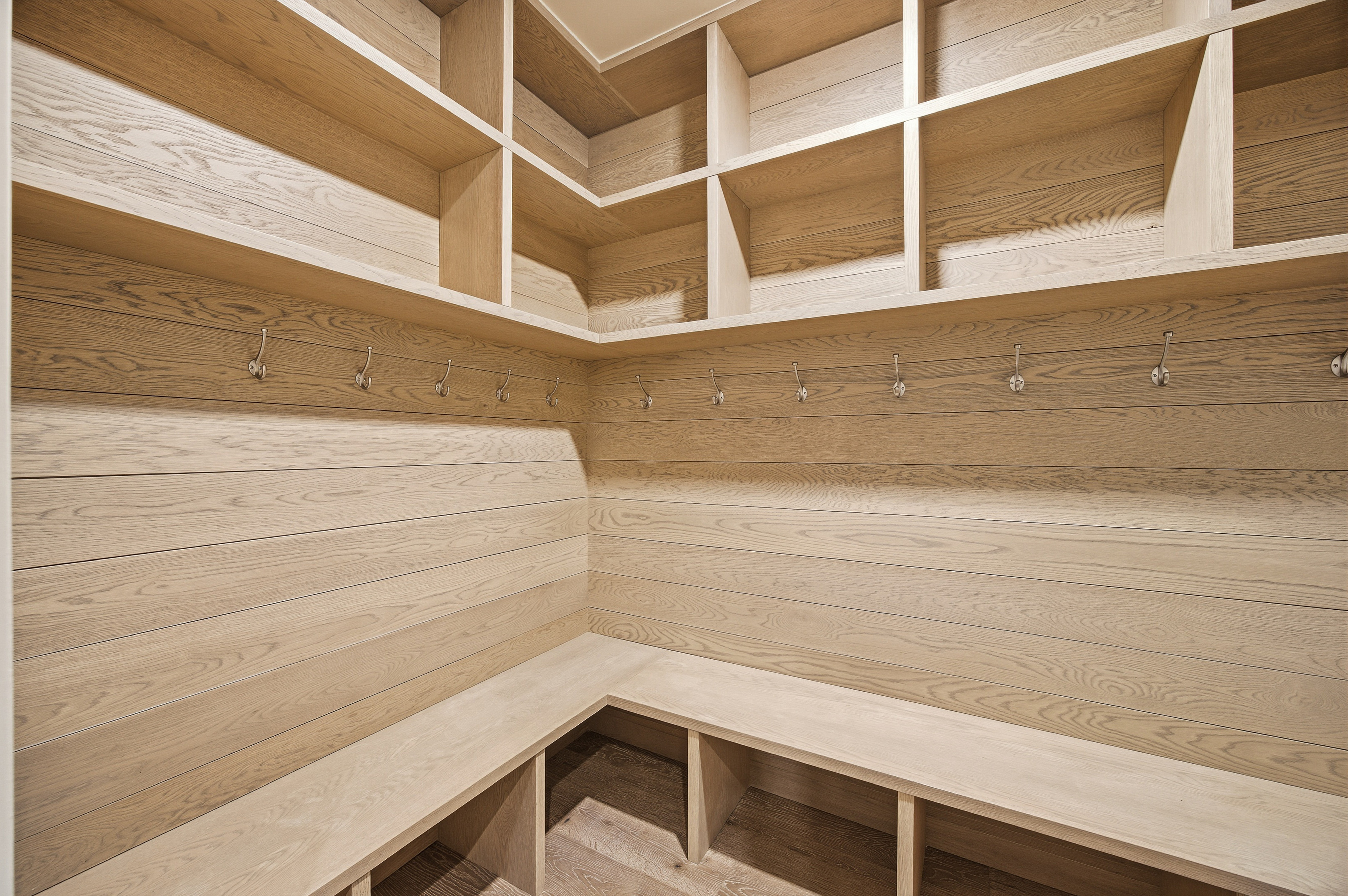 This is a well-organized walk-in closet featuring light wood shelving and paneling. The closet includes open shelves, a bench with storage underneath, and a row of hooks for hanging items. The design maximizes storage space and creates a clean, modern aesthetic.